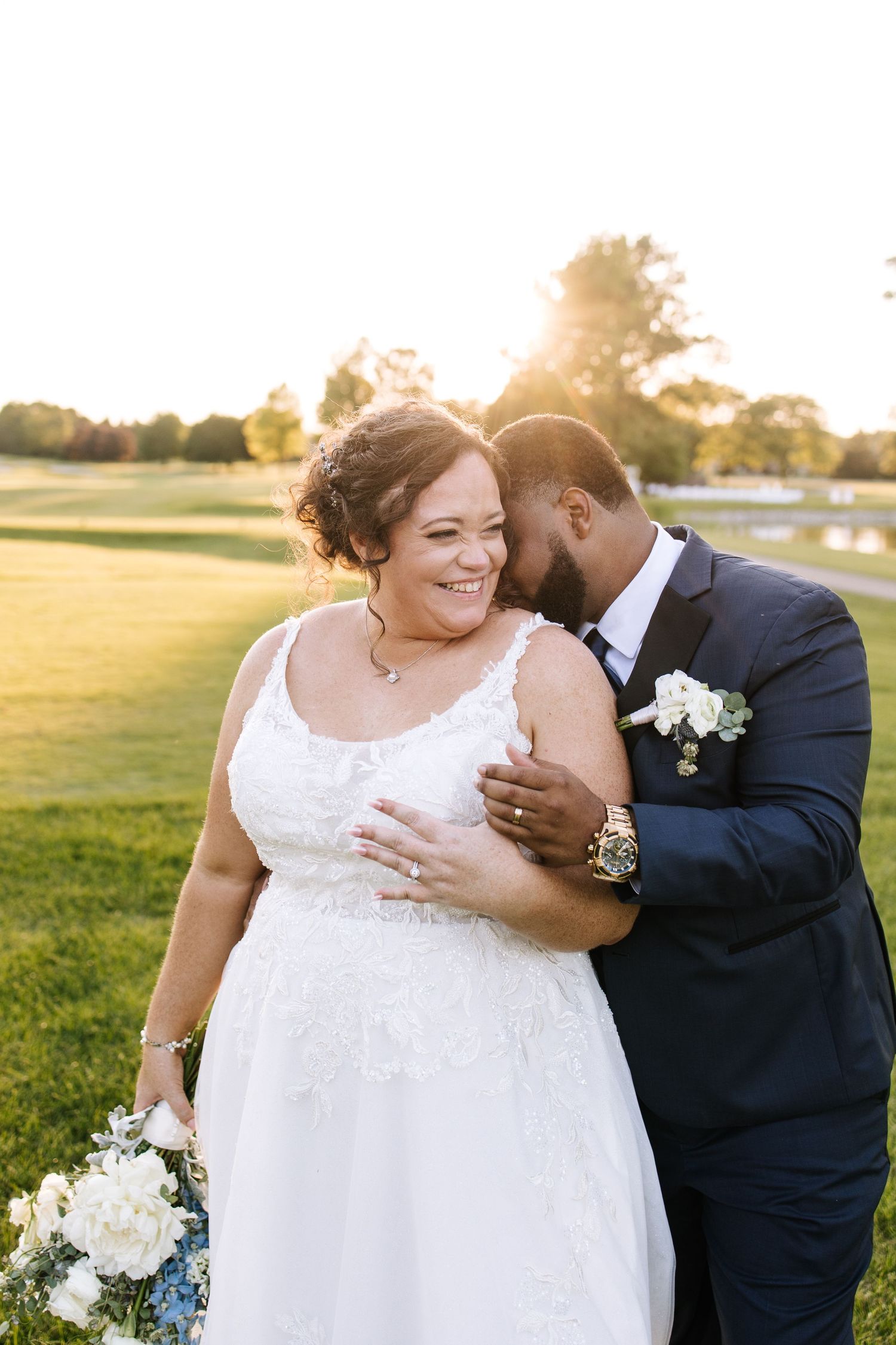 A romantic wedding photoshoot in a sunlit field with a couple in white dress and dark suit embracing.
