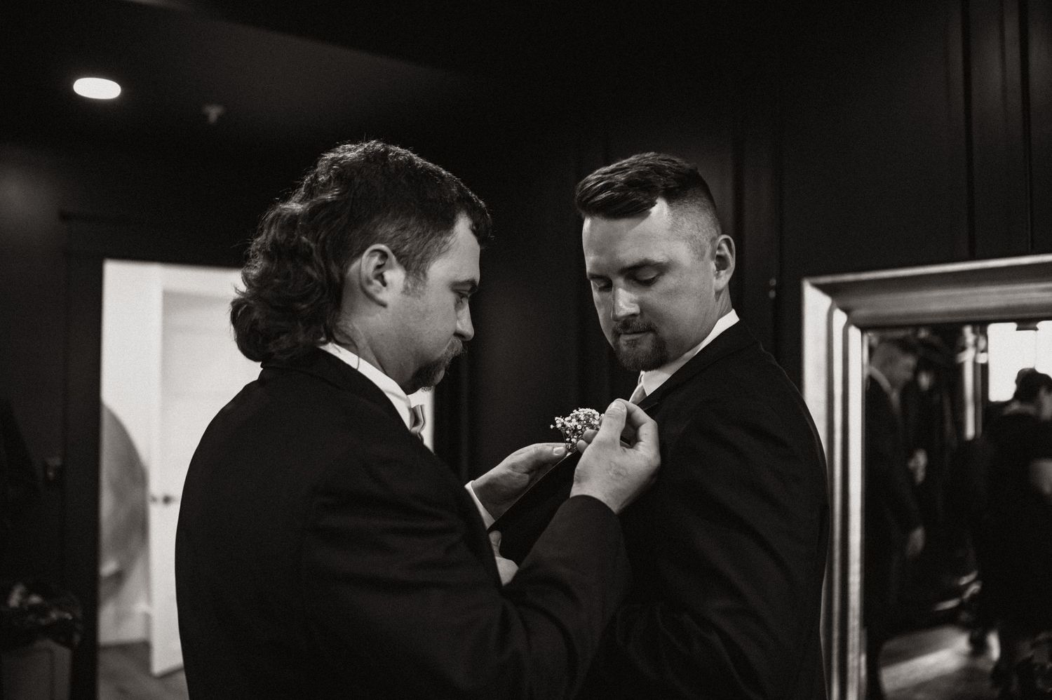 Two people helping each other with formal attire preparations in dark, moody lighting.