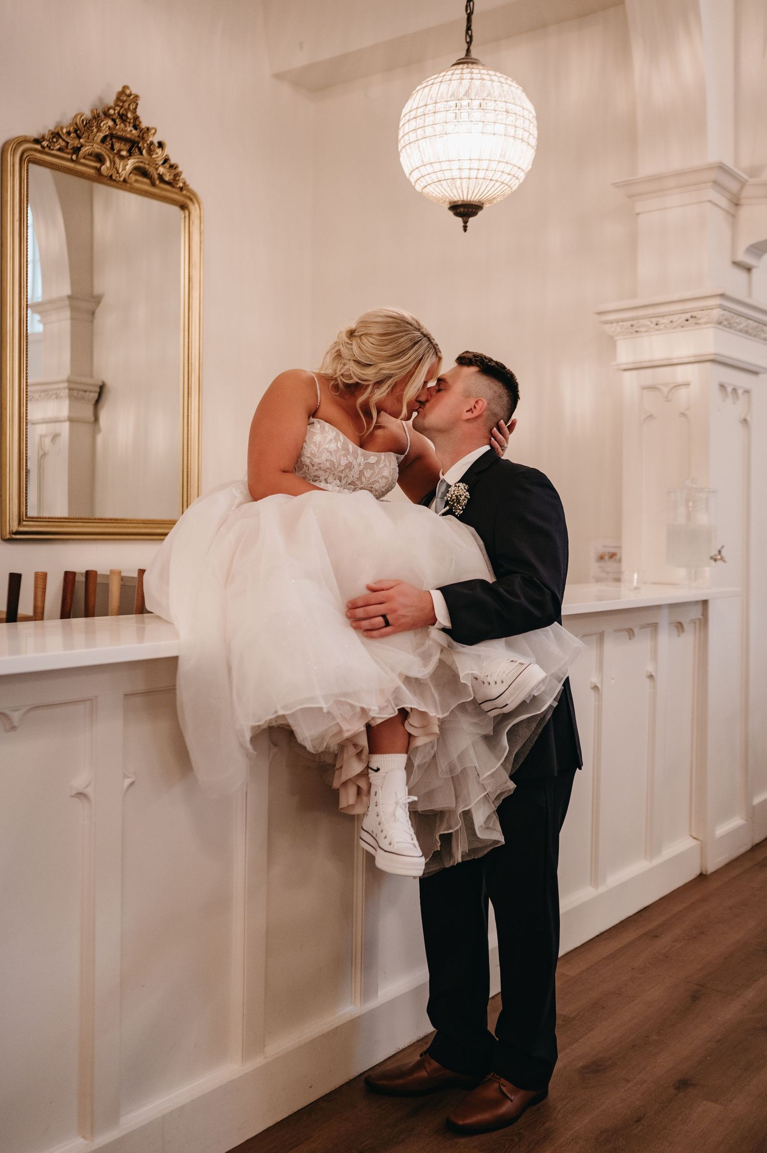 Romantic wedding moment captured under a crystal globe light fixture with ornate gold mirror.