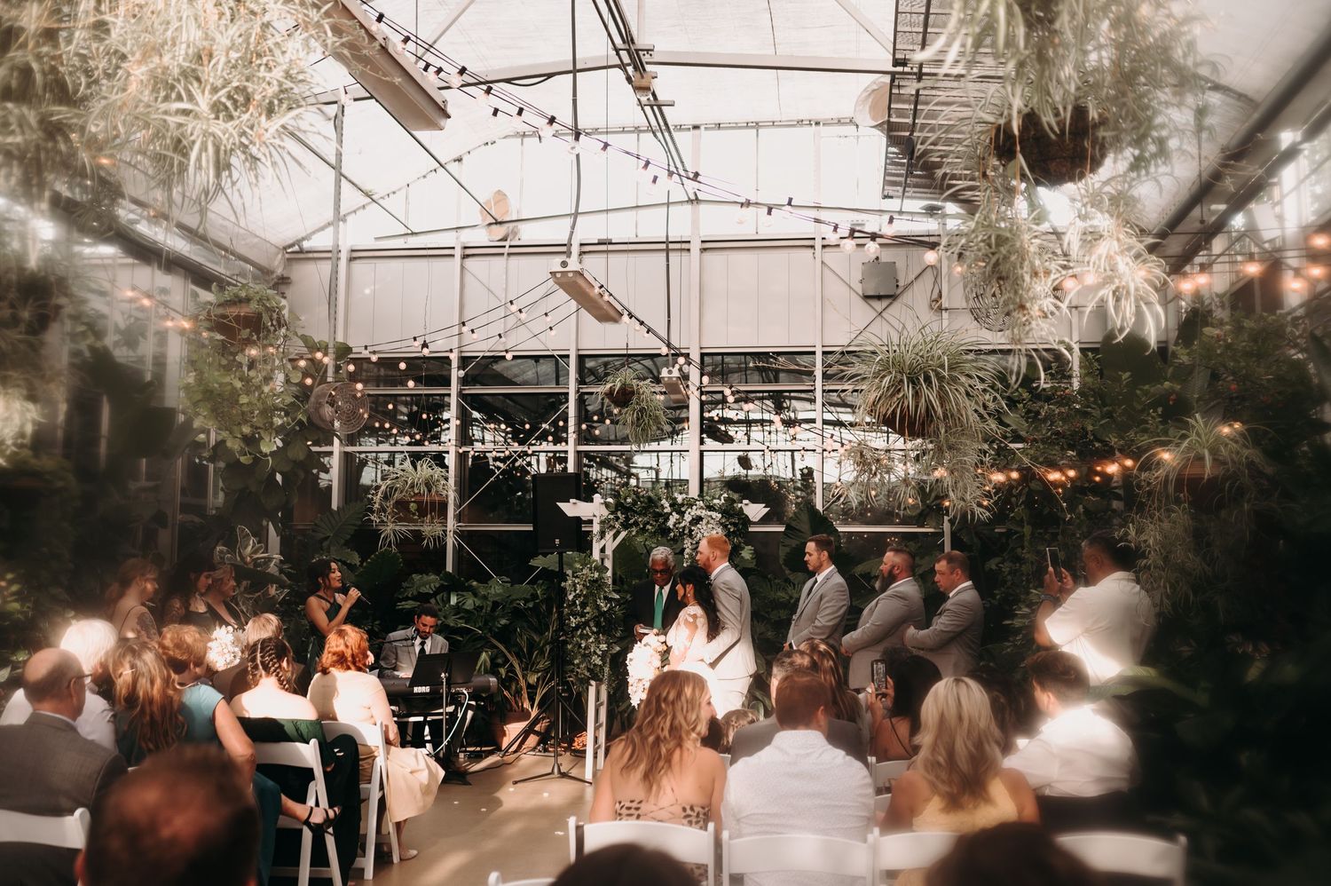 Wedding celebration in a glass-enclosed greenhouse with hanging lights and natural decor.