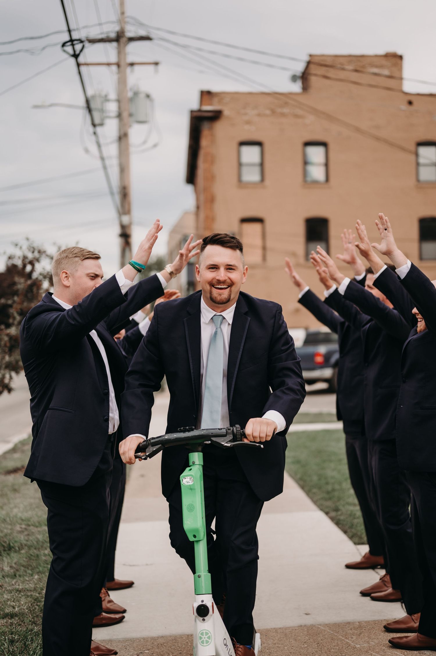 Groomsman having fun on electric scooter while others celebrate outside brick building.