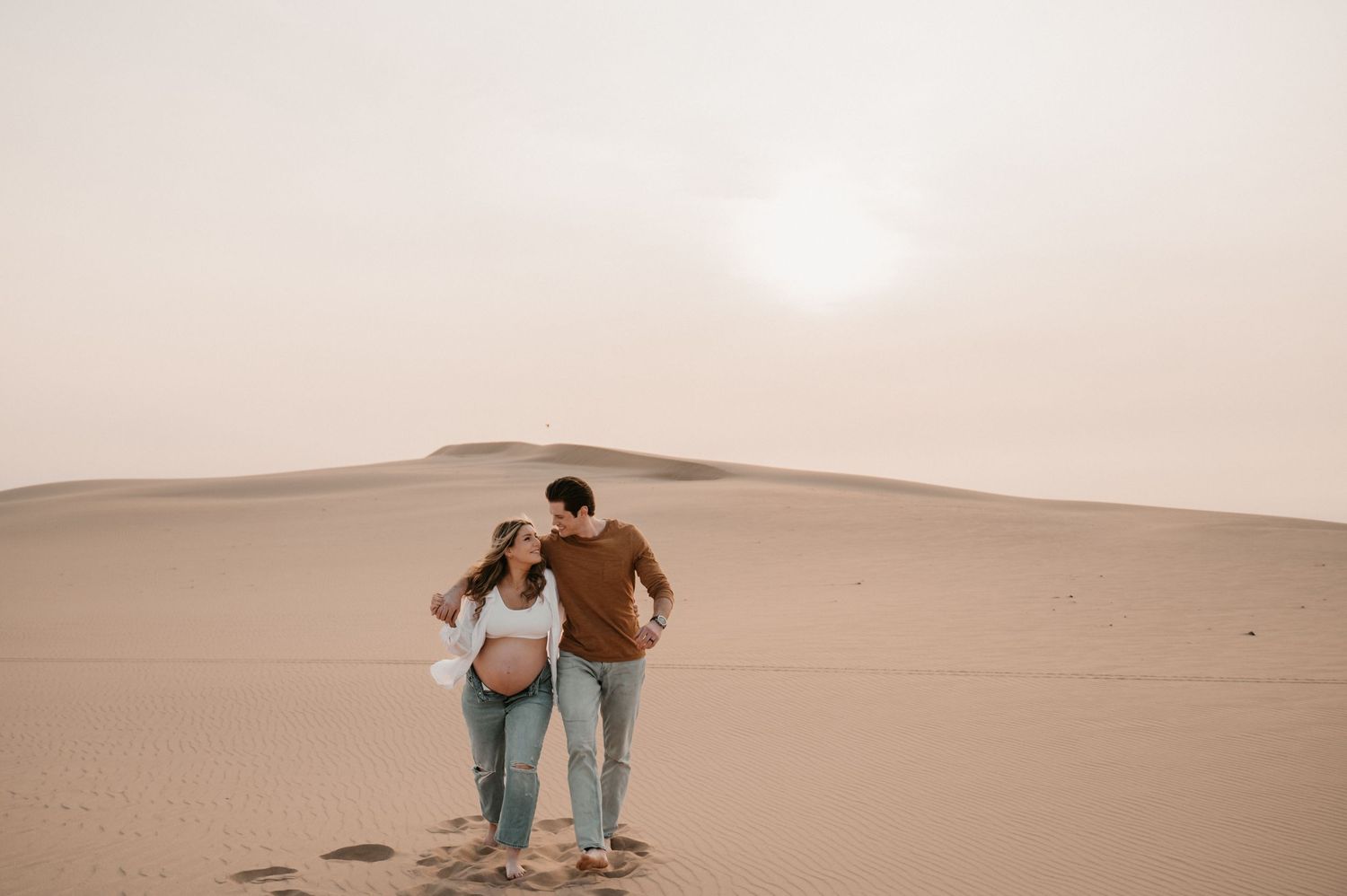 A couple embraces while standing on desert sand dunes during sunset.