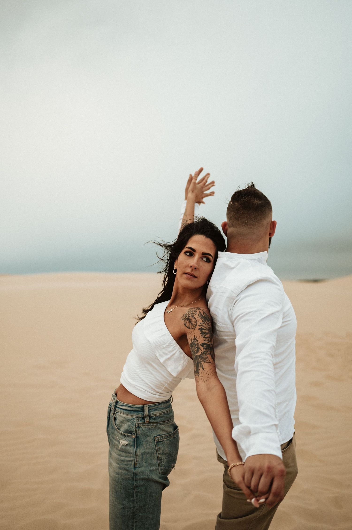 A couple in casual attire strikes a dramatic pose on desert sand dunes.