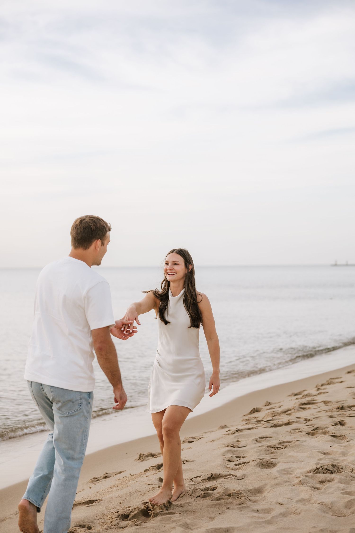 Joyful couple holding hands and walking along the shoreline, woman in white dress smiling at her partner.