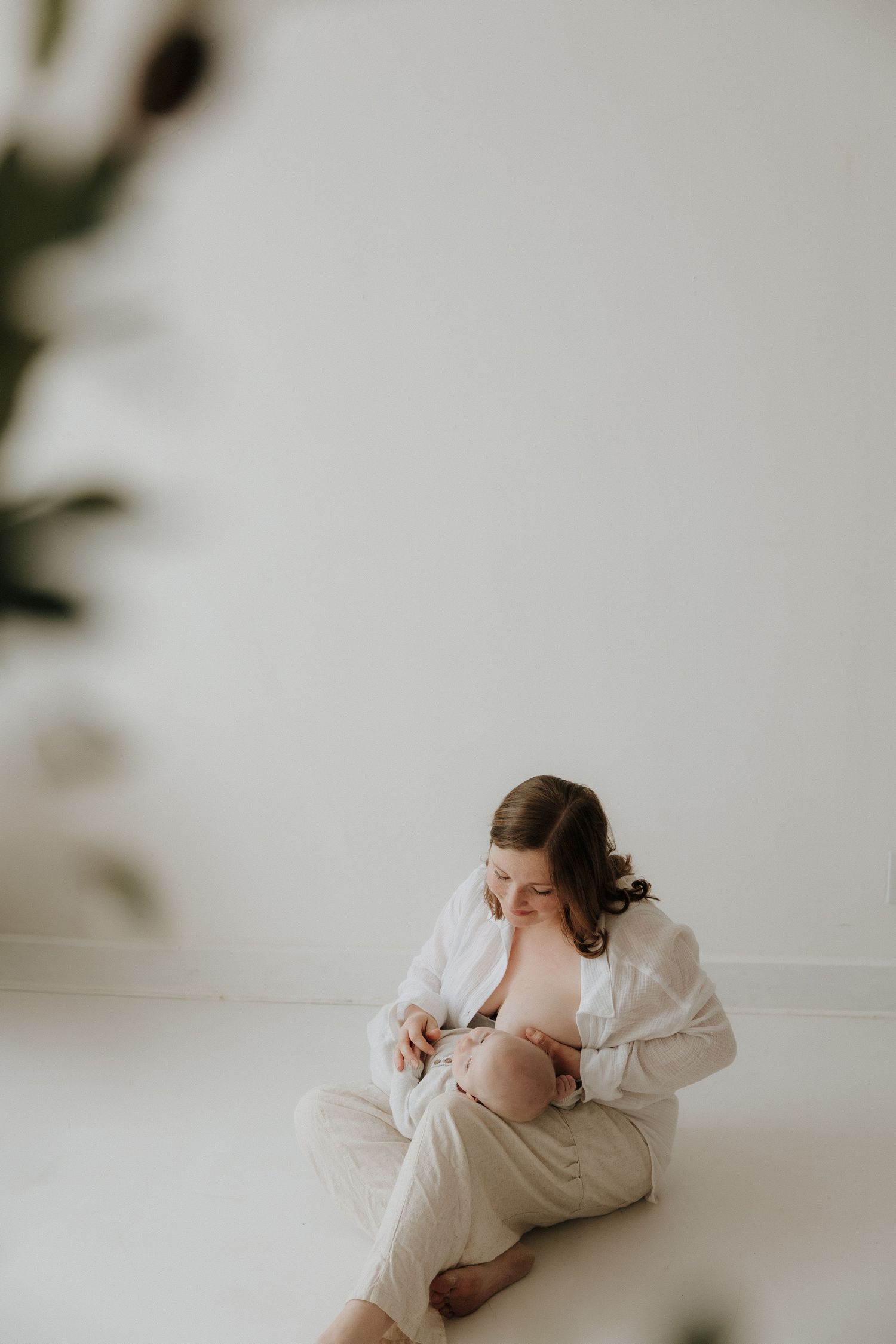 A mother sits on the floor breastfeeding her newborn baby, wearing a white shirt in a bright minimalist room.