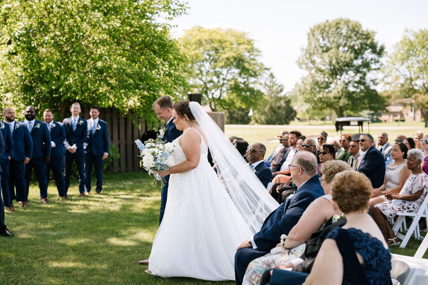 Outdoor wedding ceremony setup with guests seated in white chairs under trees.