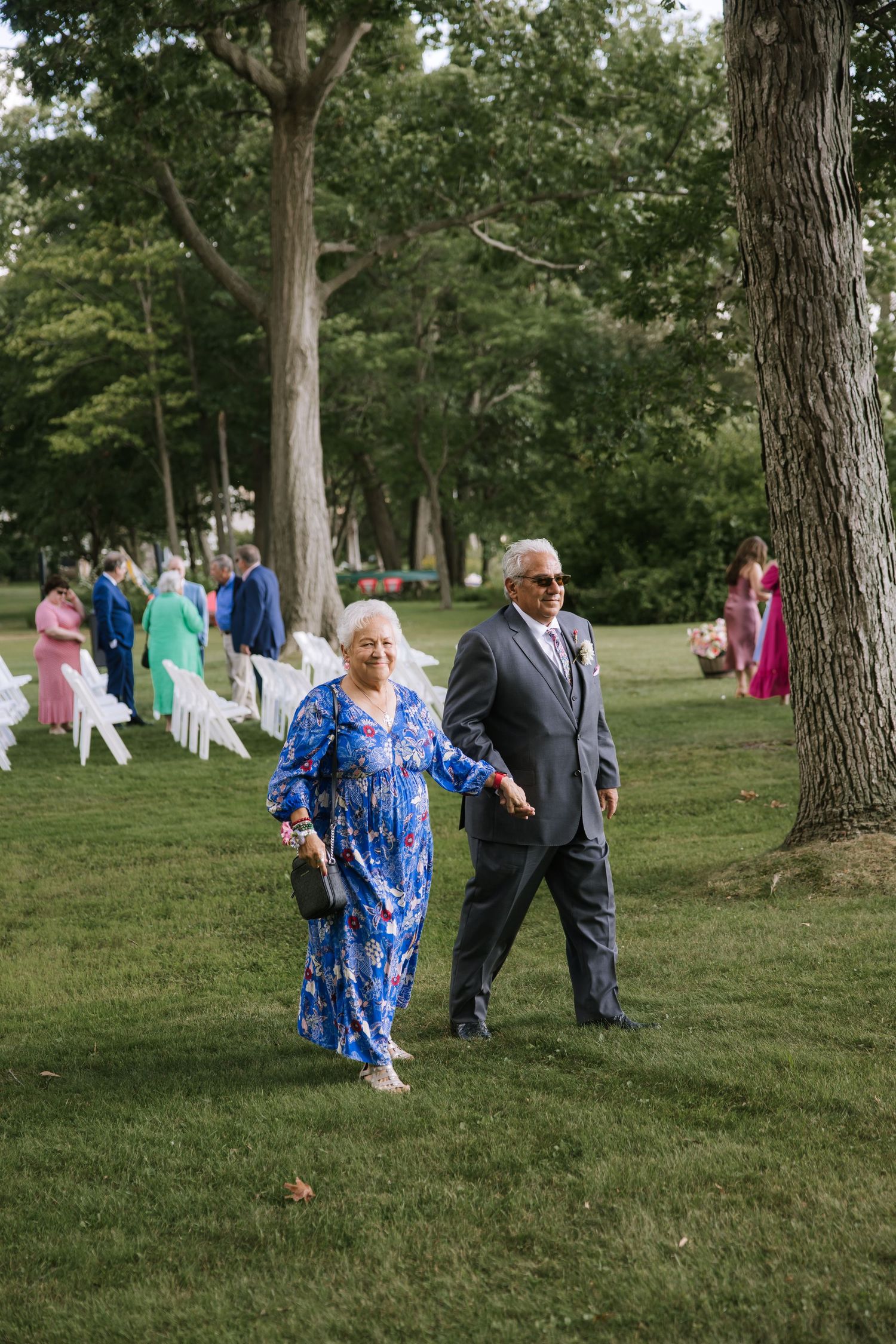 Wedding guests in formal attire mingling on a grassy lawn surrounded by trees.
