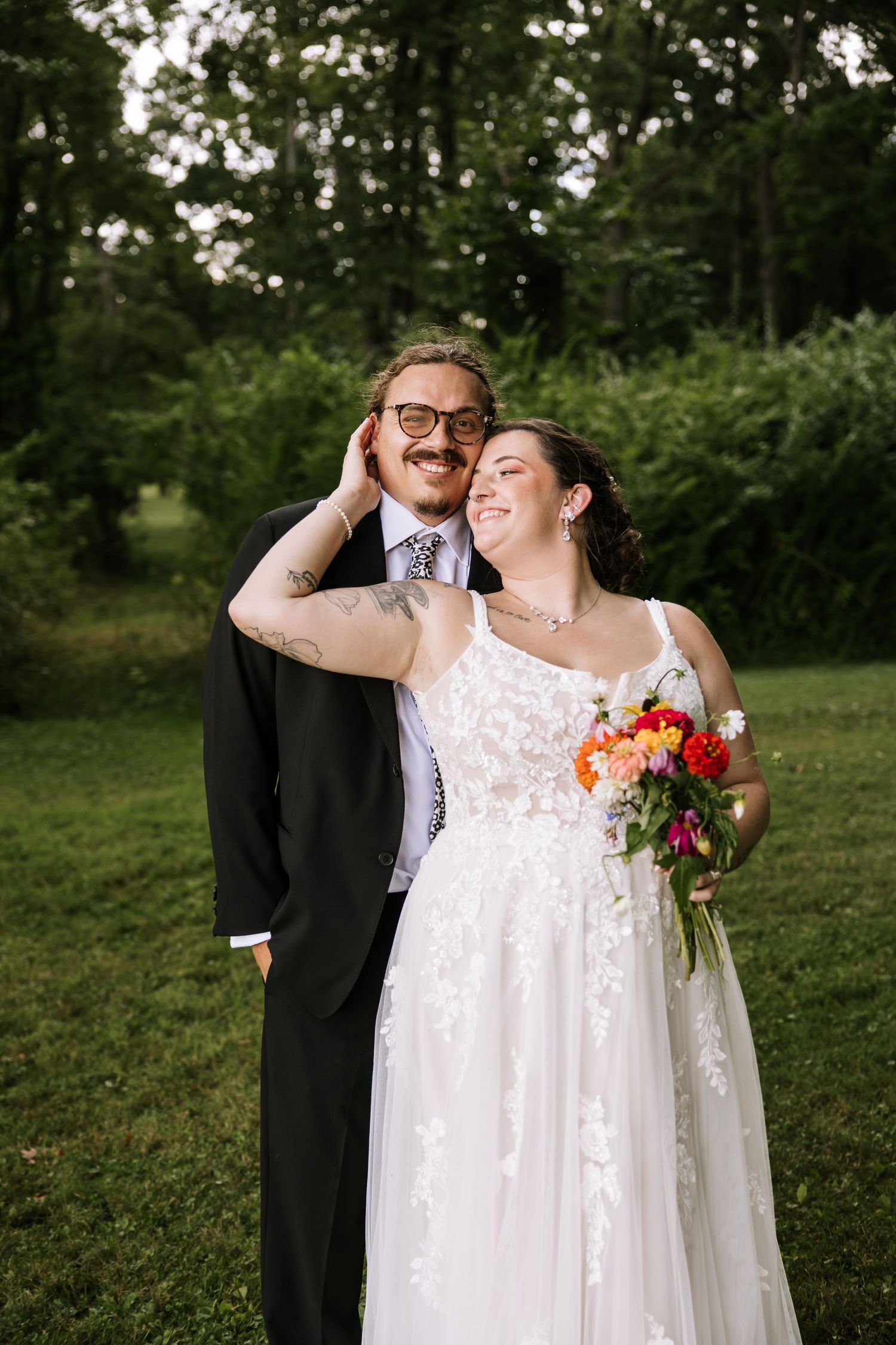 A joyful wedding couple embraces and laughs together in a wooded outdoor setting.