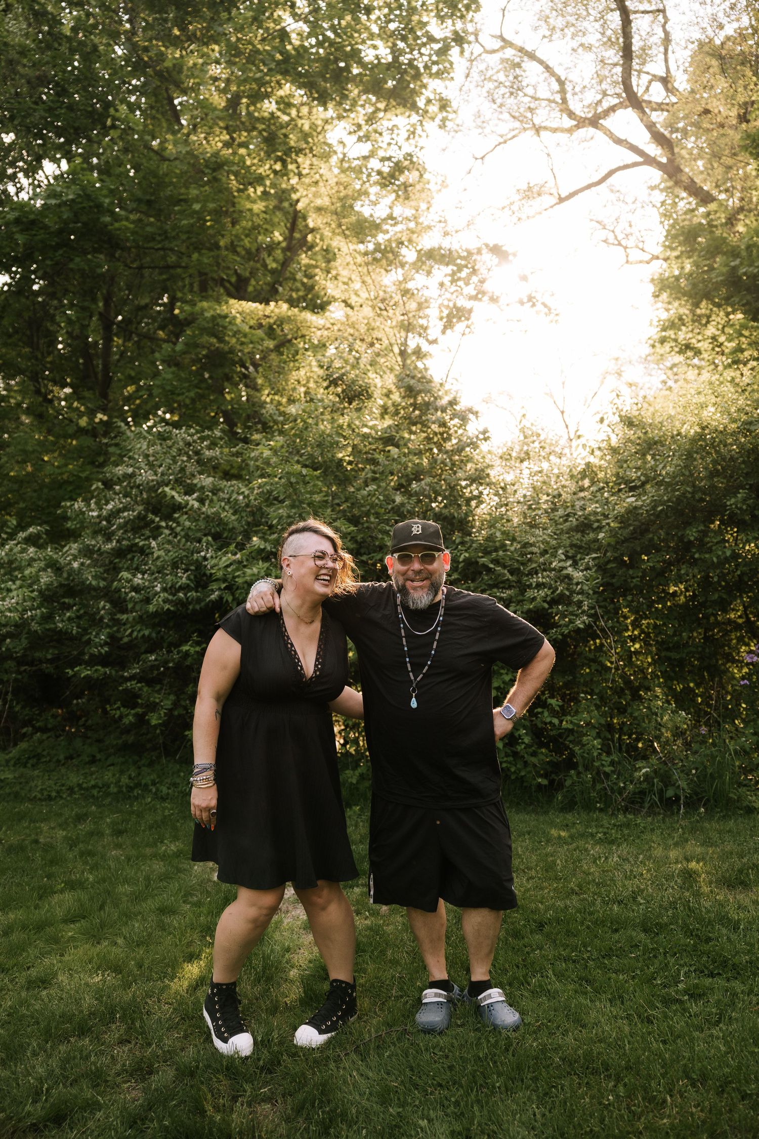 Smiling couple in black outfits standing together on grass with sunlit trees behind them.