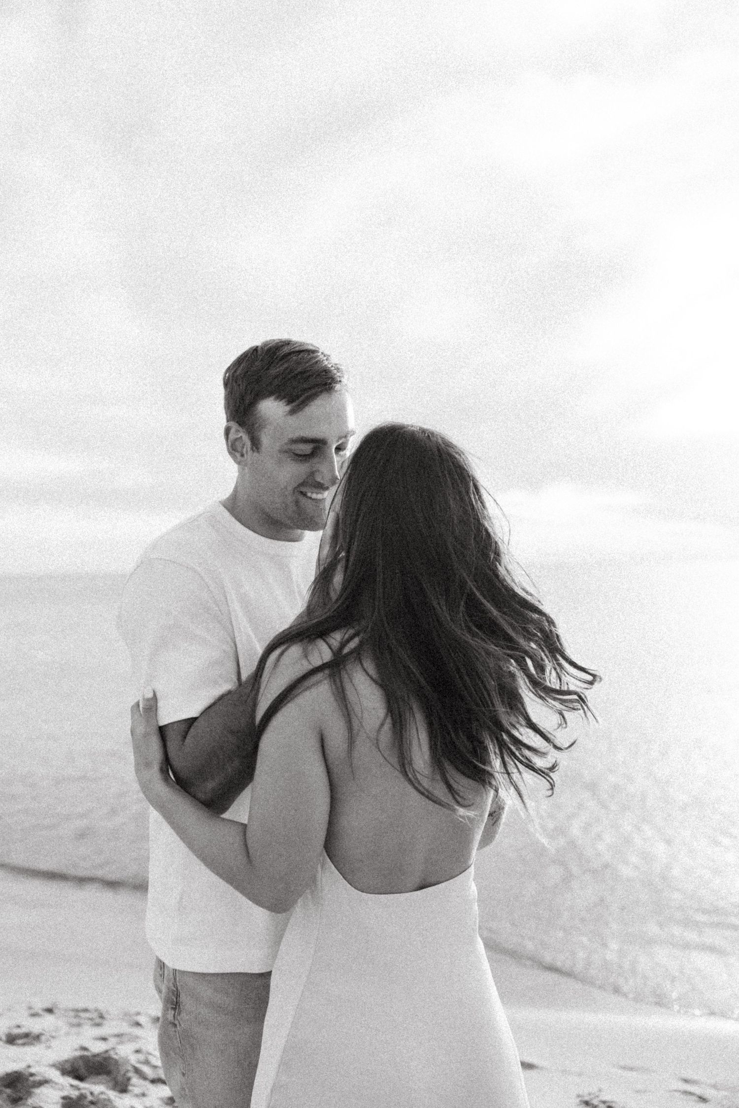 Black and white beach photo of smiling man embracing woman, her hair blowing in wind, ocean backdrop.
