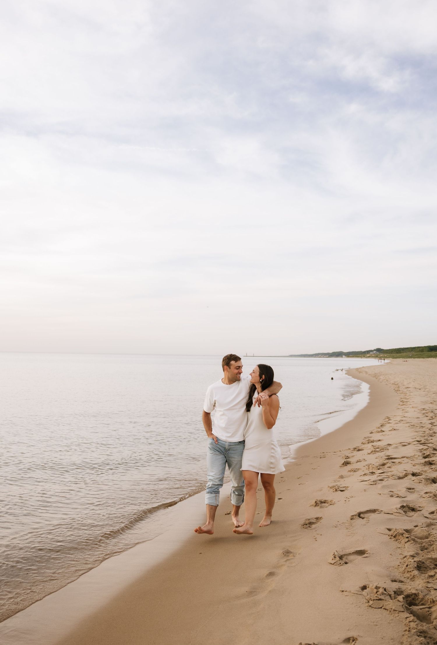 Couple walking barefoot on sandy beach shoreline, man in white tee and jeans, woman in white dress, calm ocean backdrop.