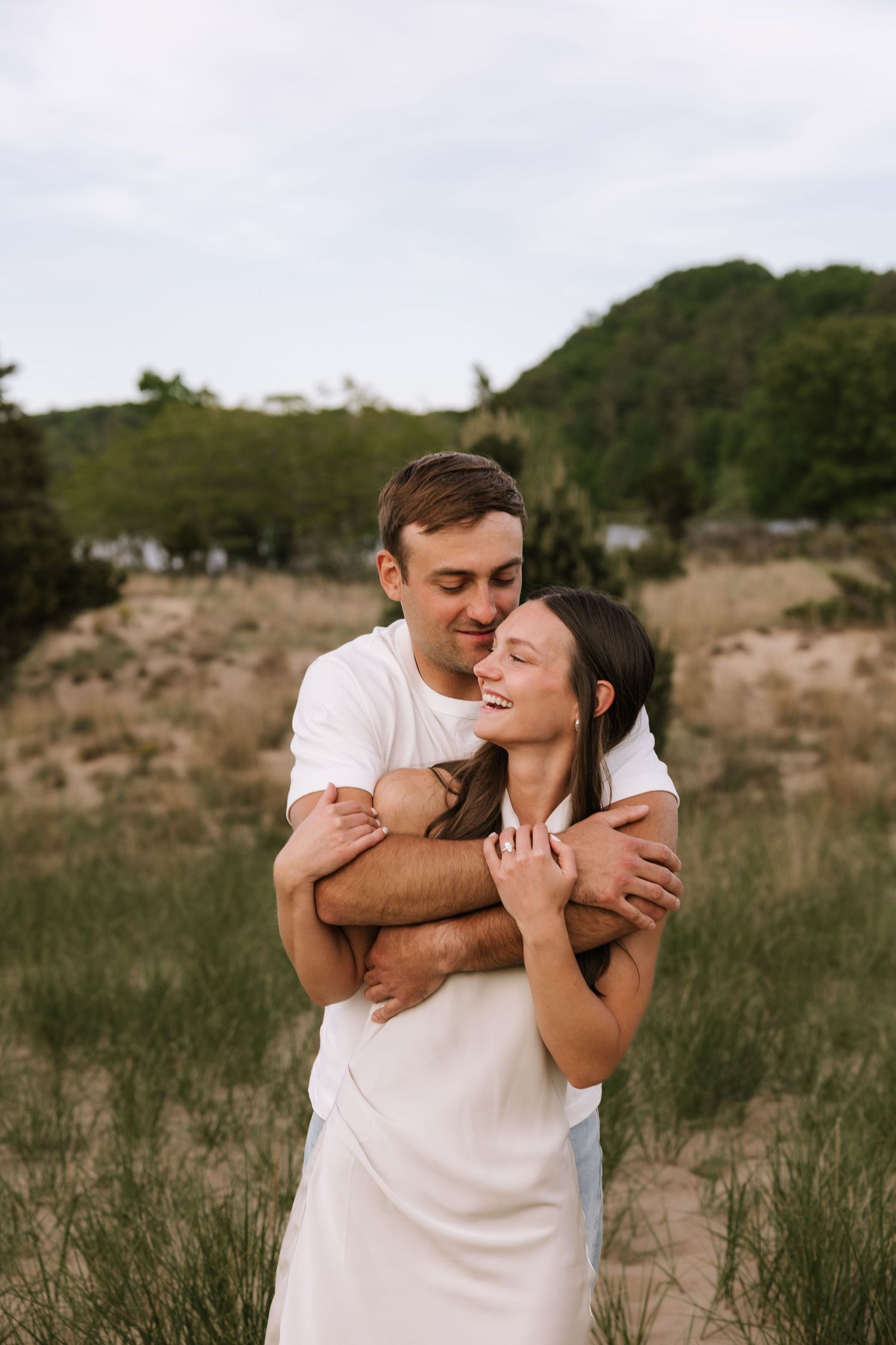 Man hugging woman from behind on sandy beach, both smiling, wearing white outfits, green hills in background.