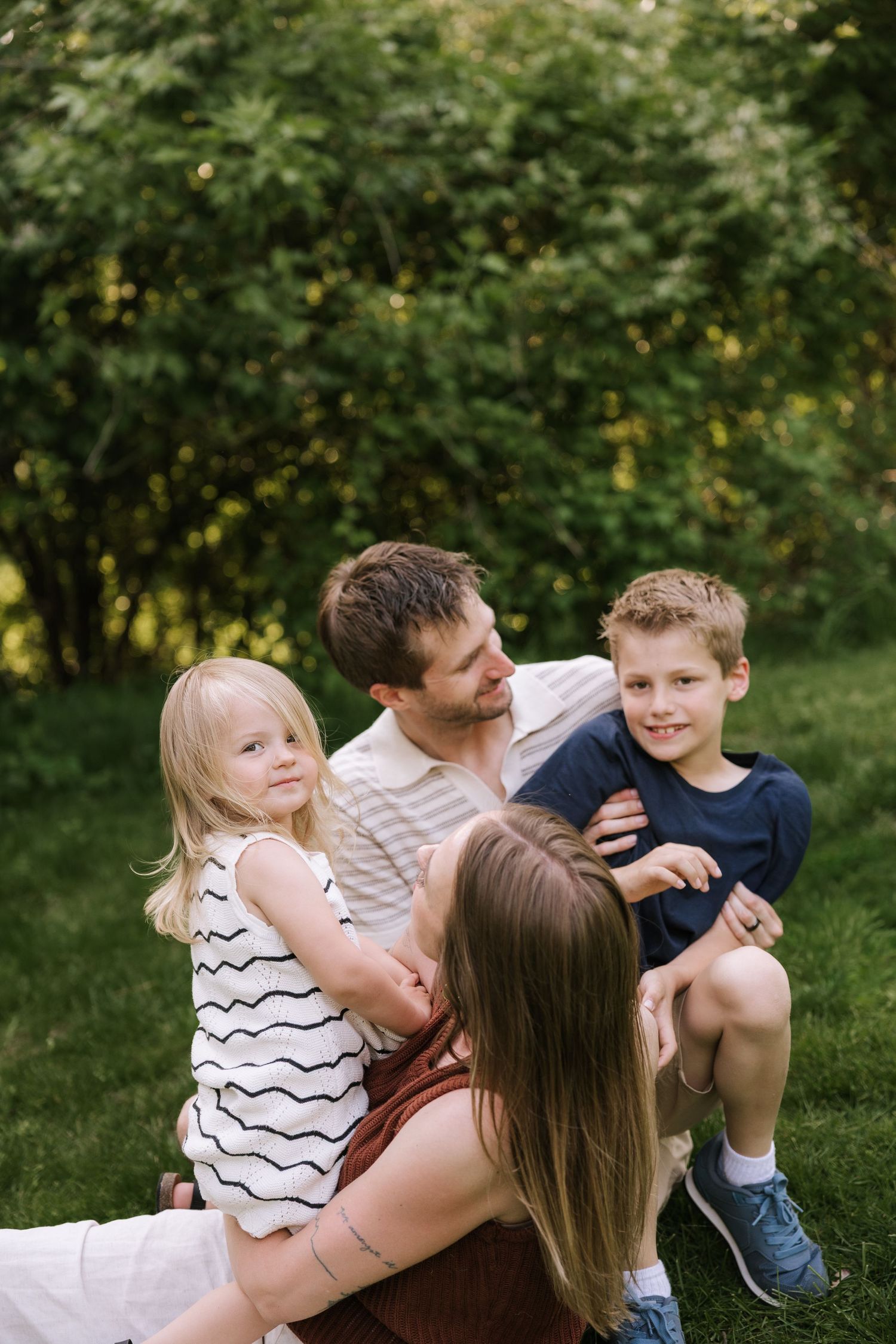 Family of four sitting together on grass in green garden, parents bonding with young children.