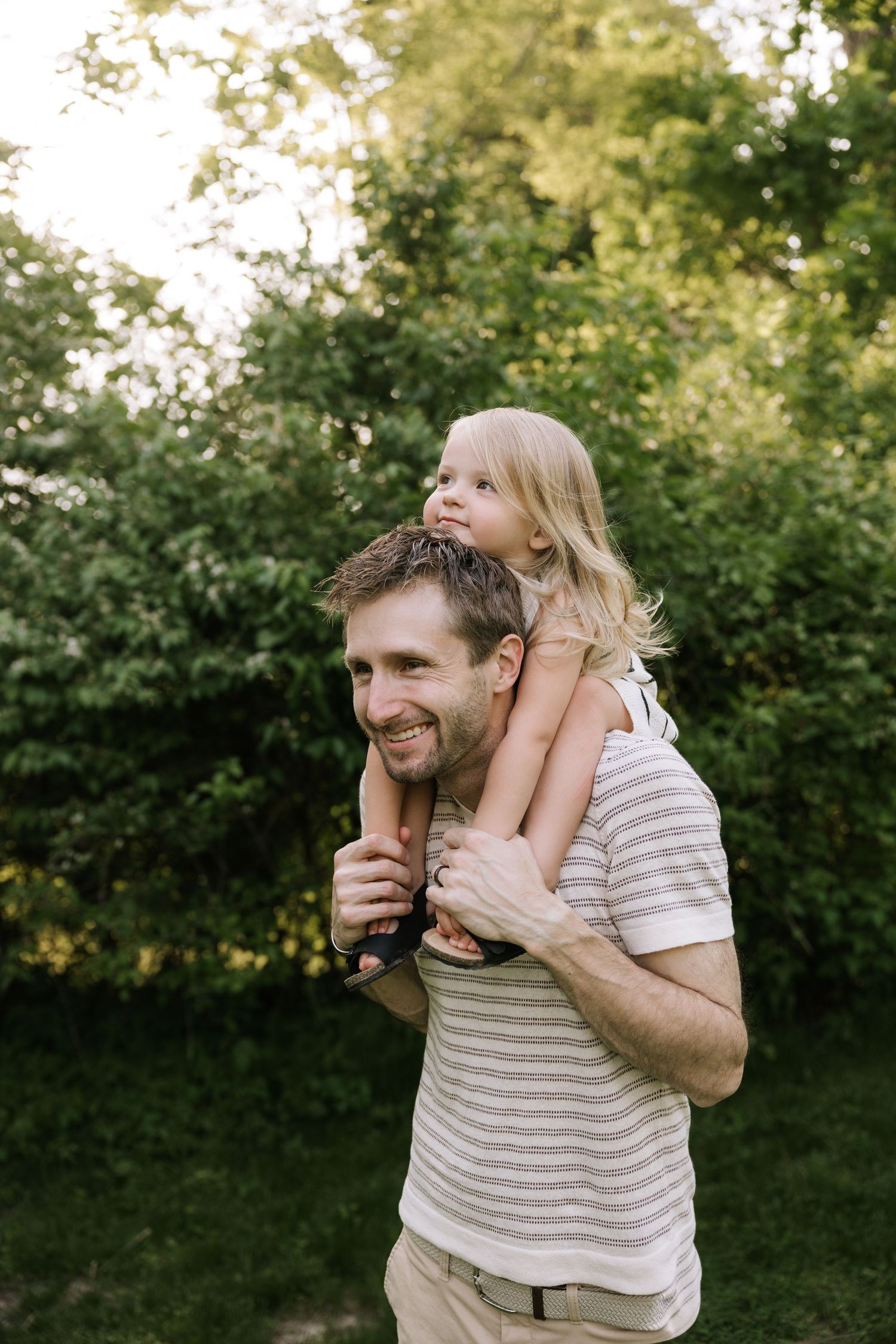 Happy father carrying blonde child on shoulders in sunny green backyard.