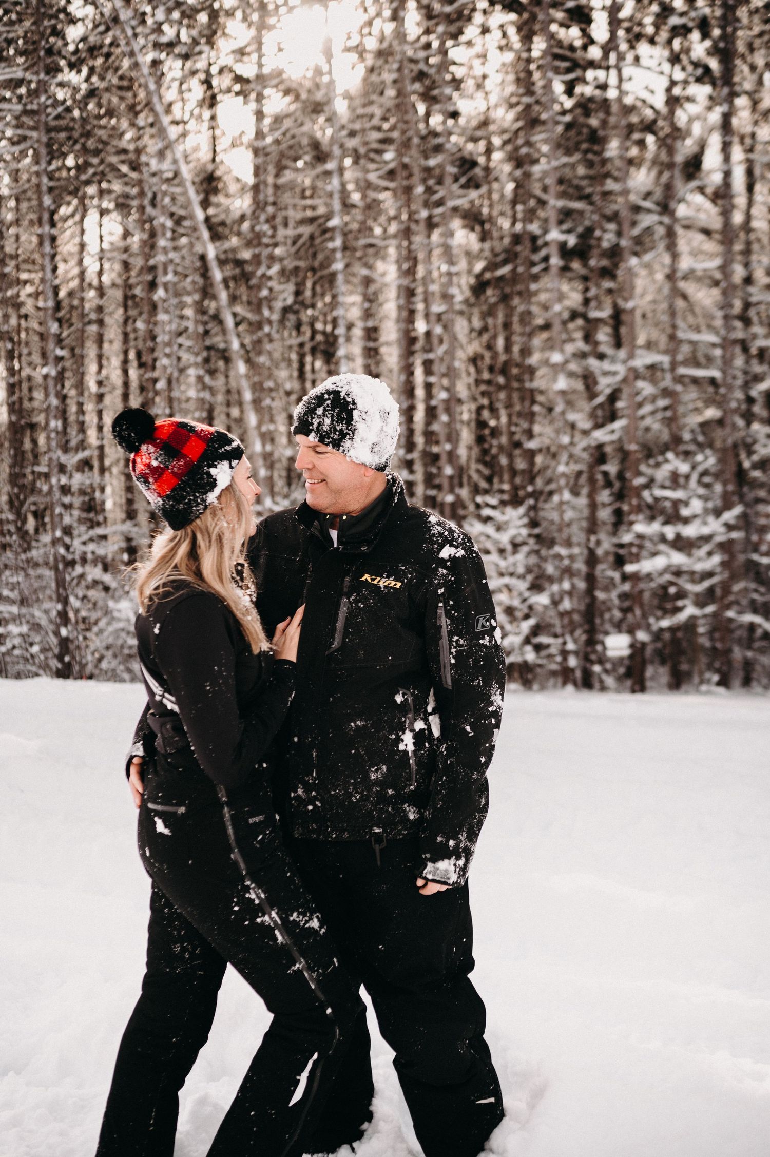 Two people in winter gear share an intimate moment against a backdrop of snow-covered pine trees.
