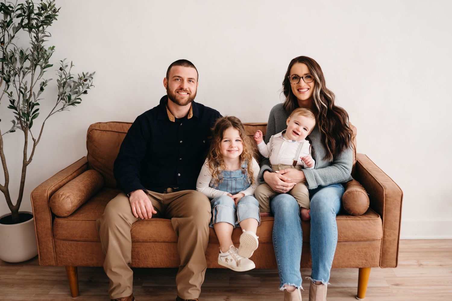 A family sits together on a brown leather couch against a white wall with a decorative plant in the corner.