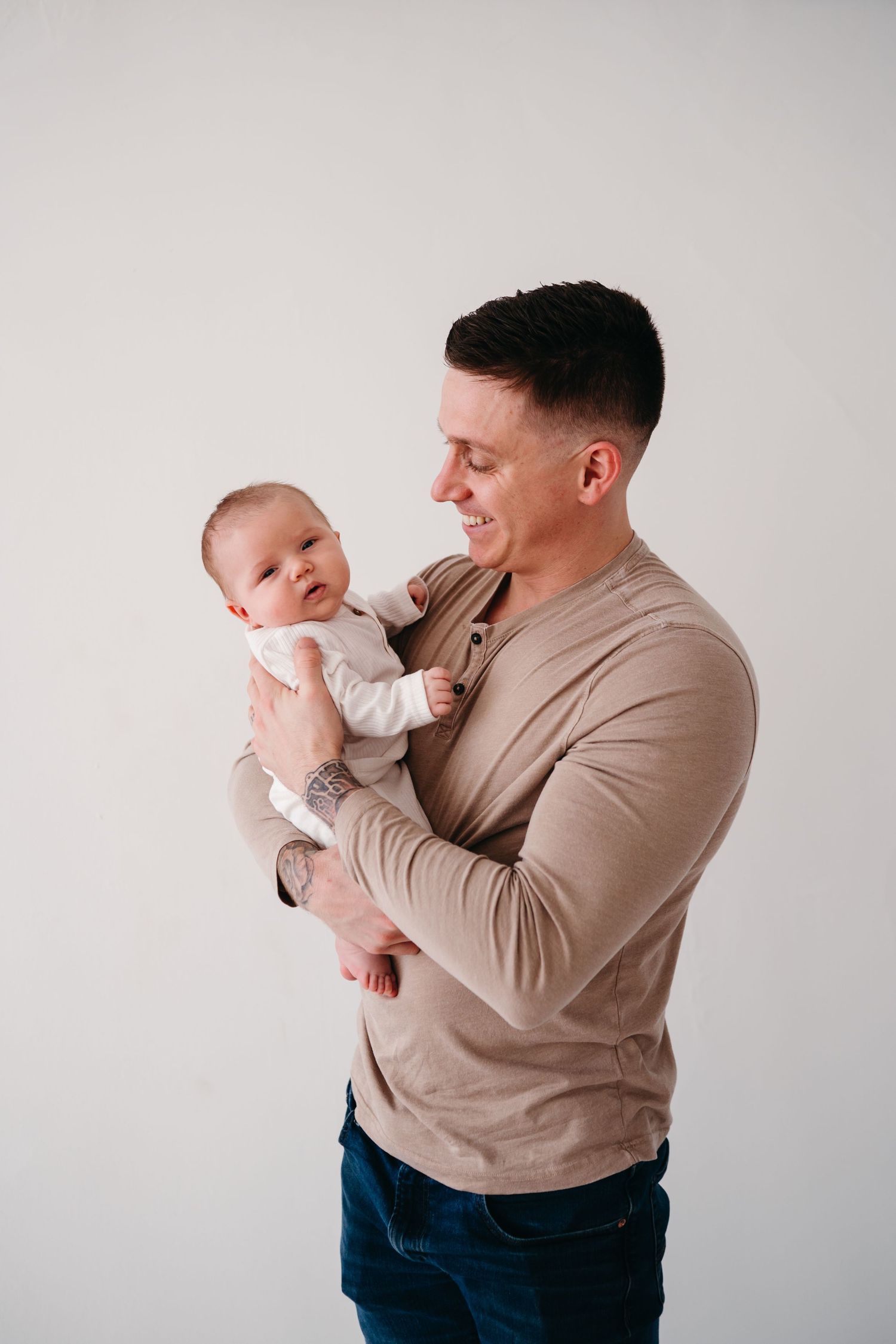 Adult in beige sweater holds and smiles at happy baby wearing white outfit against light background.