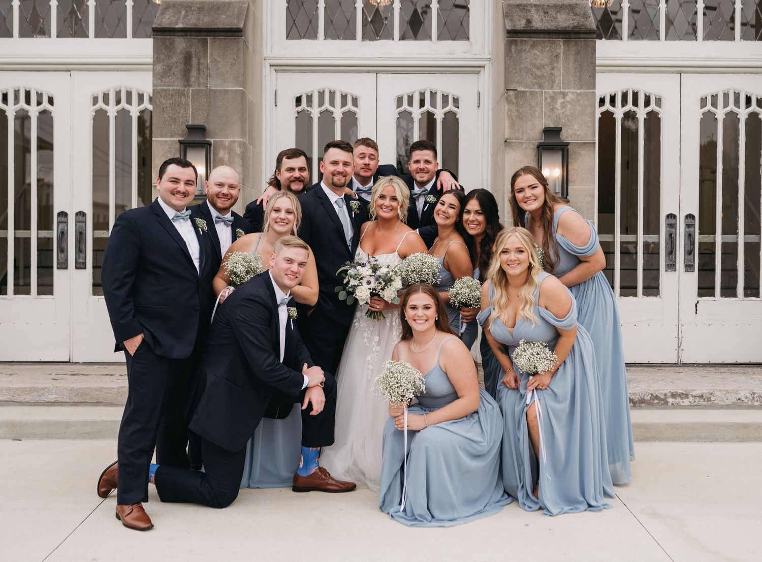 Wedding party gathered for casual group photo in front of ornate church windows.