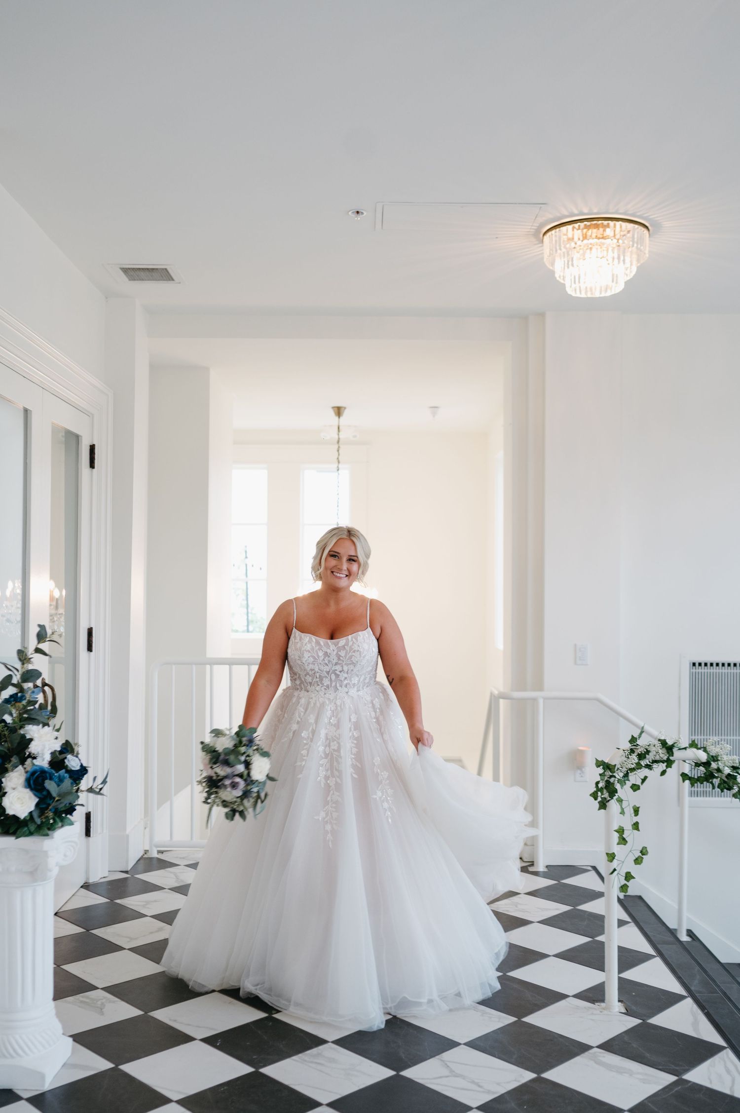 Bride twirls in flowing white wedding dress on black and white checkered floor under crystal lighting.