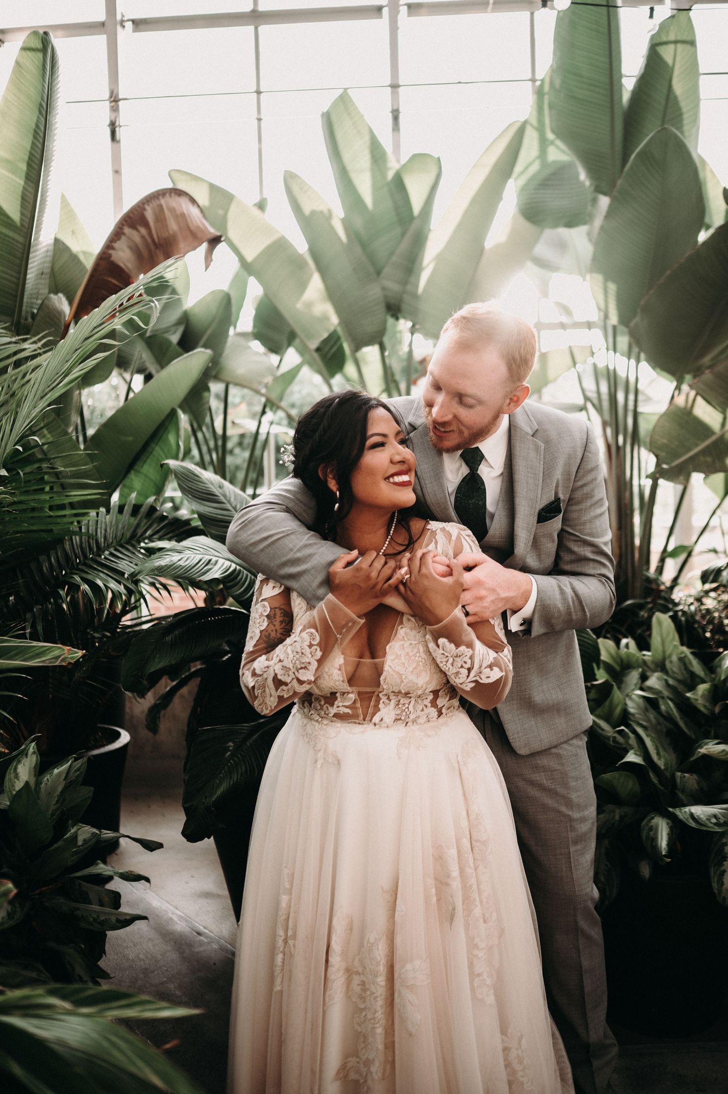 Newlyweds share a joyful moment together among tropical foliage in a greenhouse setting.