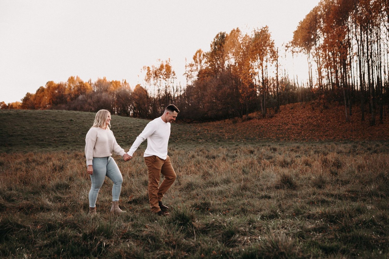 Couple walking together through rustic field with autumn trees in background.