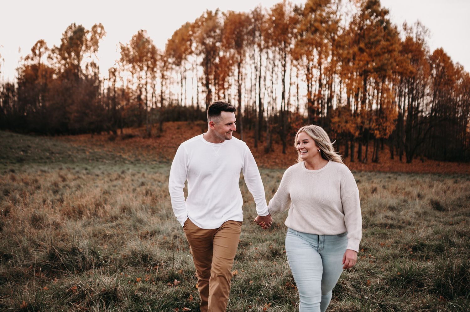 Couple walking hand in hand through an autumn field with orange trees in background.