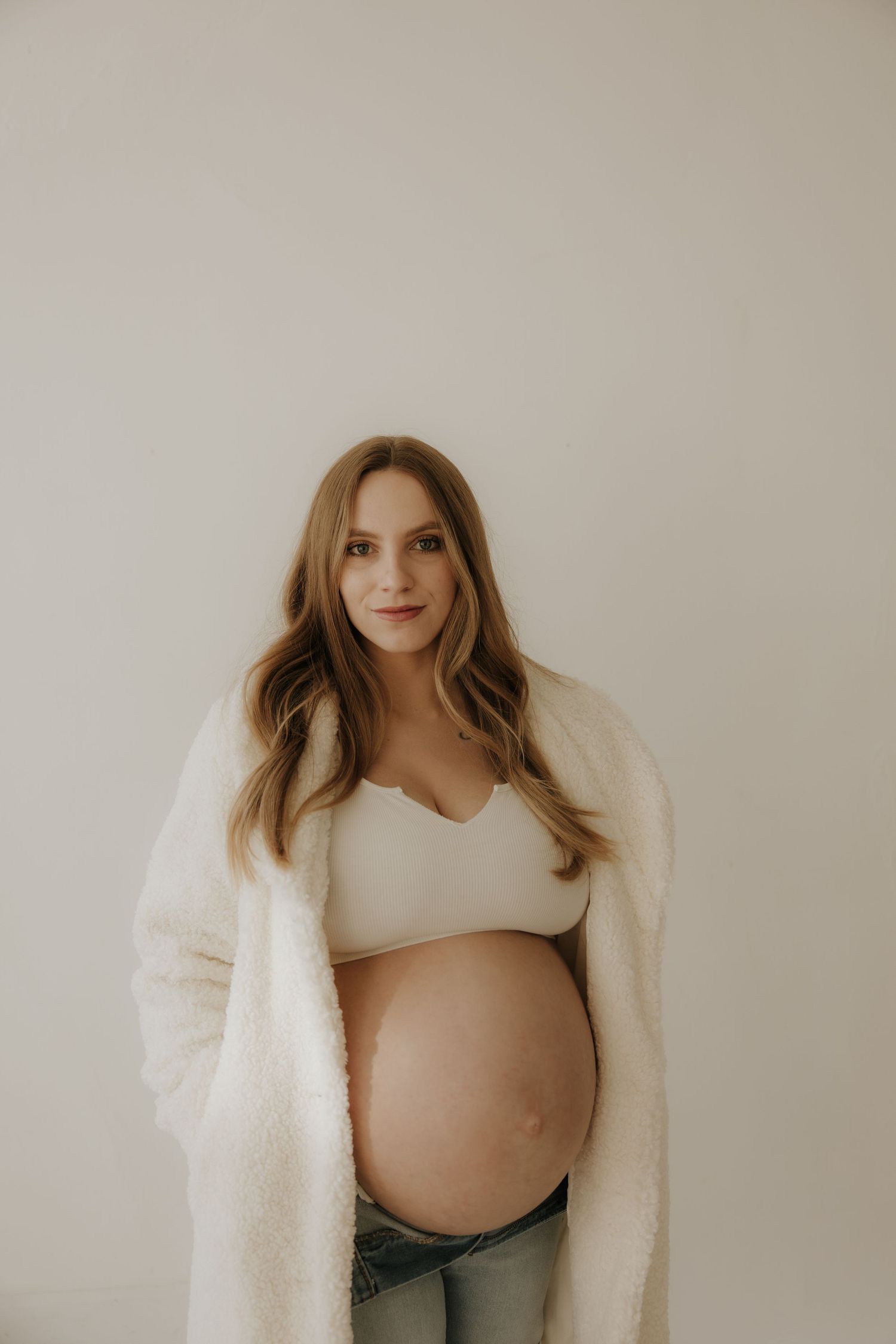 Elegant maternity session featuring woman in white bralette and fuzzy coat.
