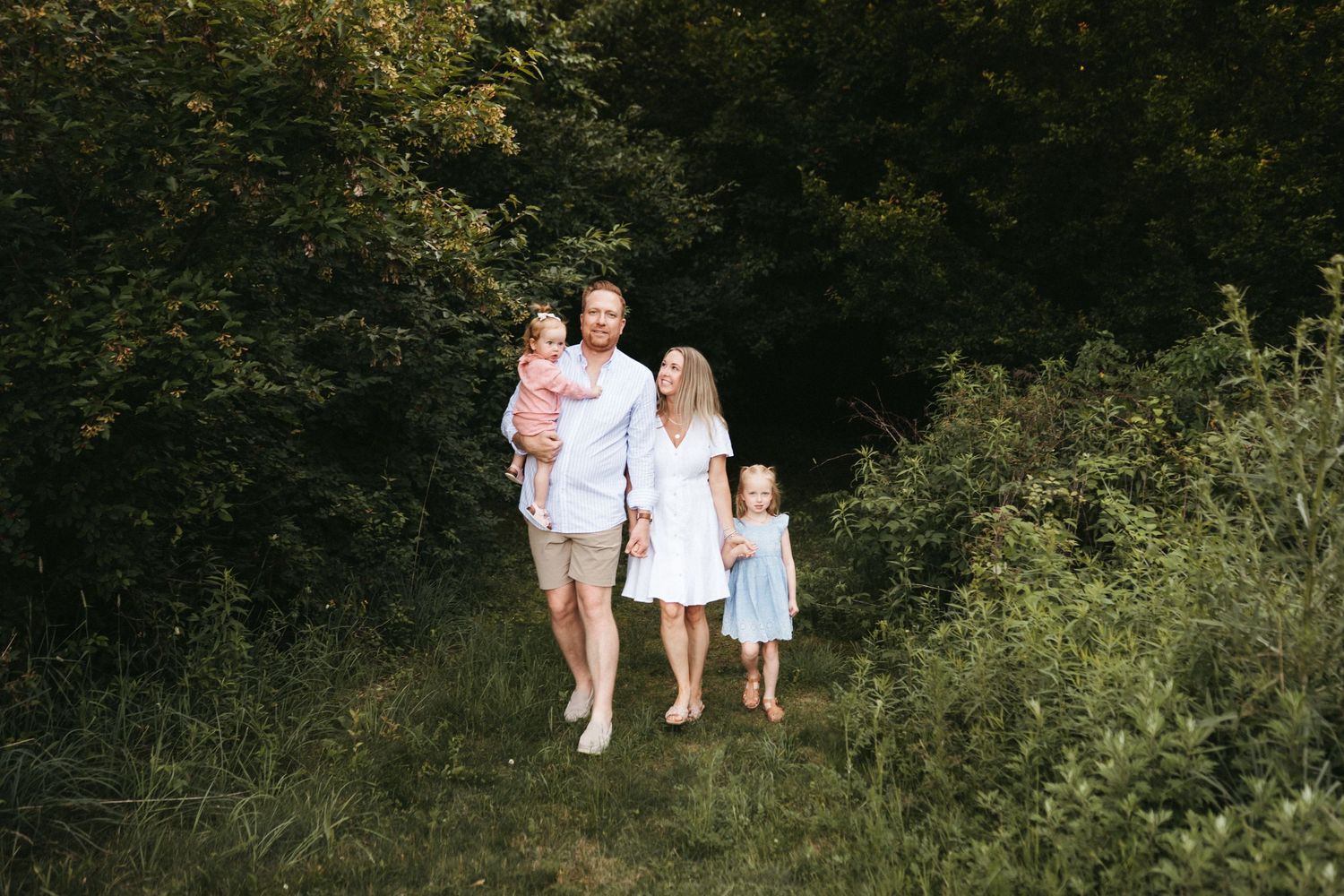 A family wearing white outfits walks together on a nature path surrounded by green foliage.
