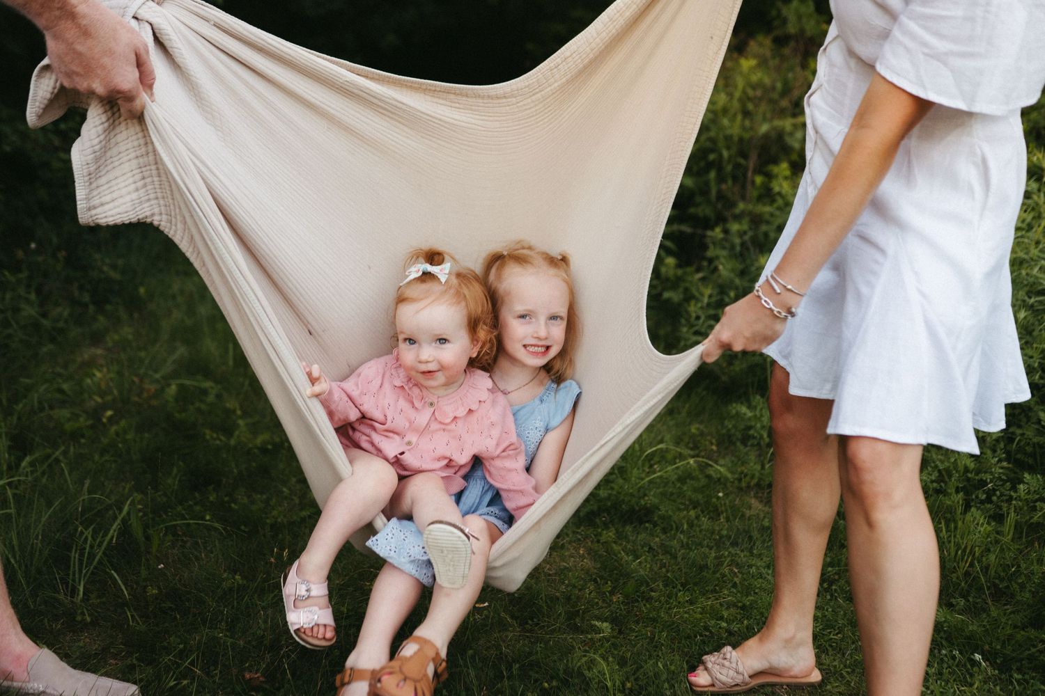 Children being playfully swung in a white blanket outdoors by adults.