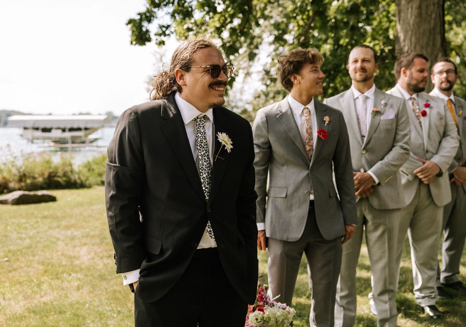 Groomsmen in gray and black suits pose together on a sunny outdoor lawn during a wedding celebration.
