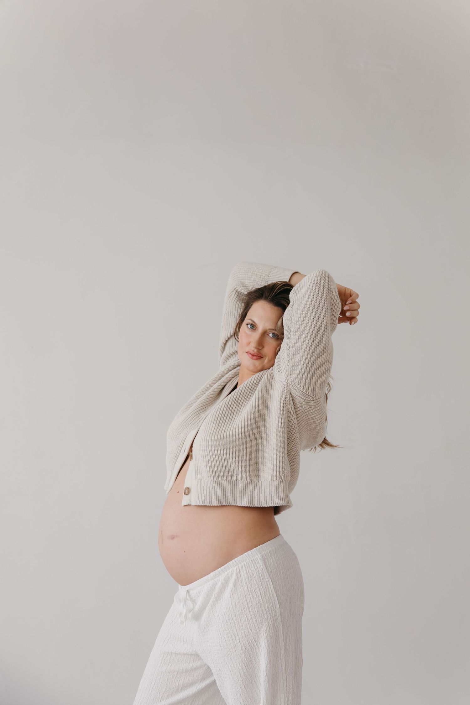 Pregnant woman in a beige ribbed knit cardigan smiles while raising her arms, exposing her baby bump against a white background.