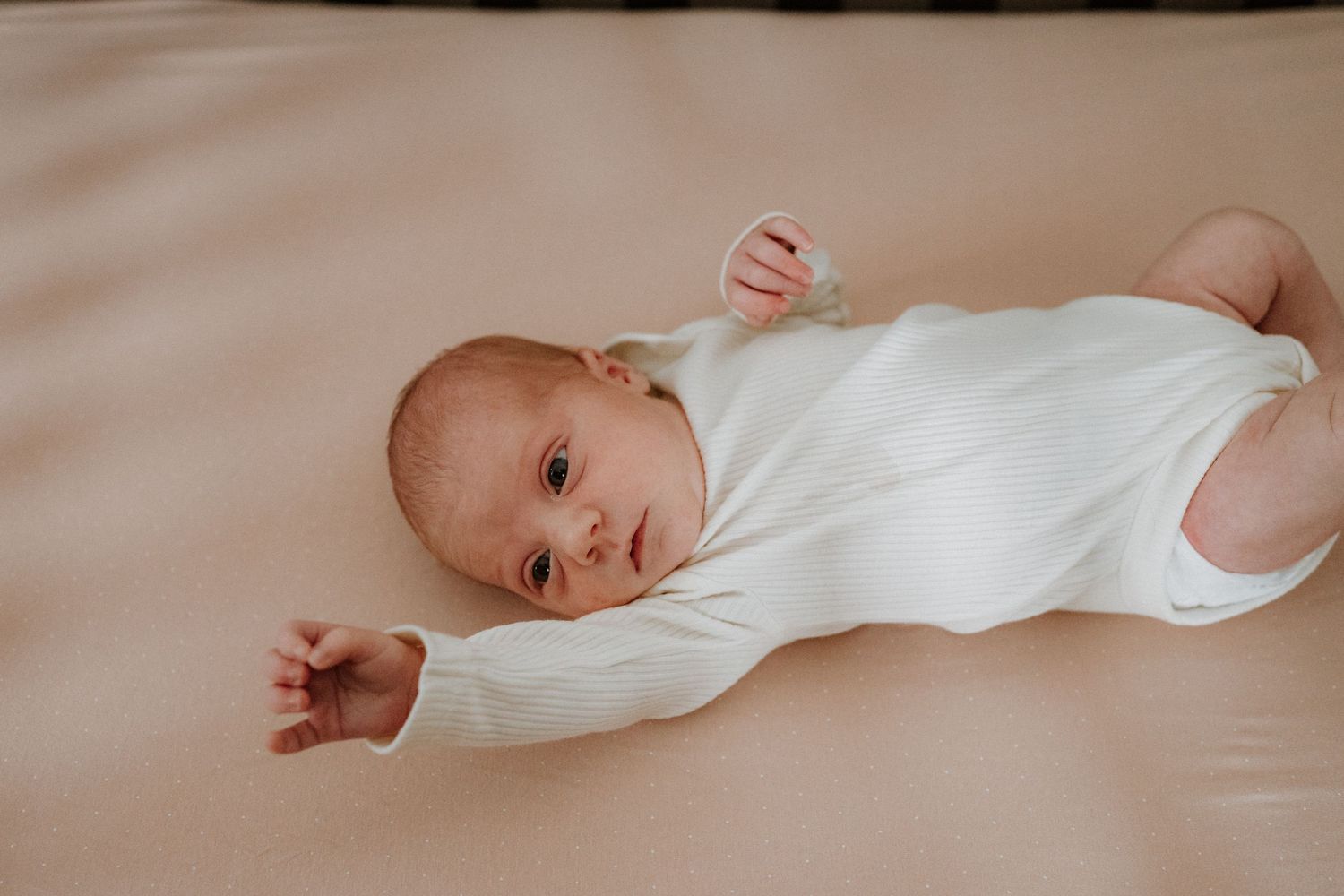 Newborn baby wearing white onesie lying on soft beige bedding during lifestyle photo session.