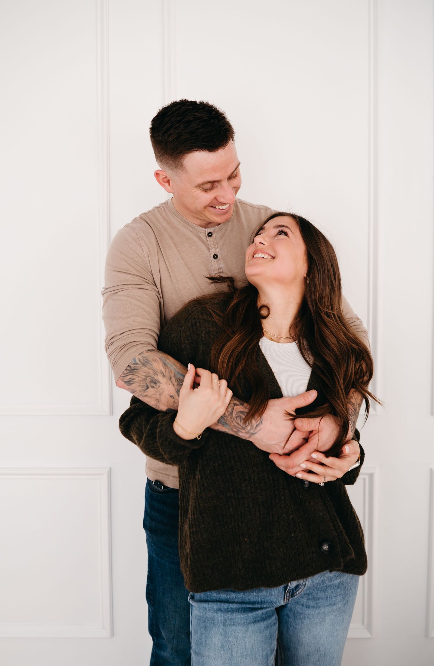 A couple shares an intimate moment while embracing and laughing against a white wall.
