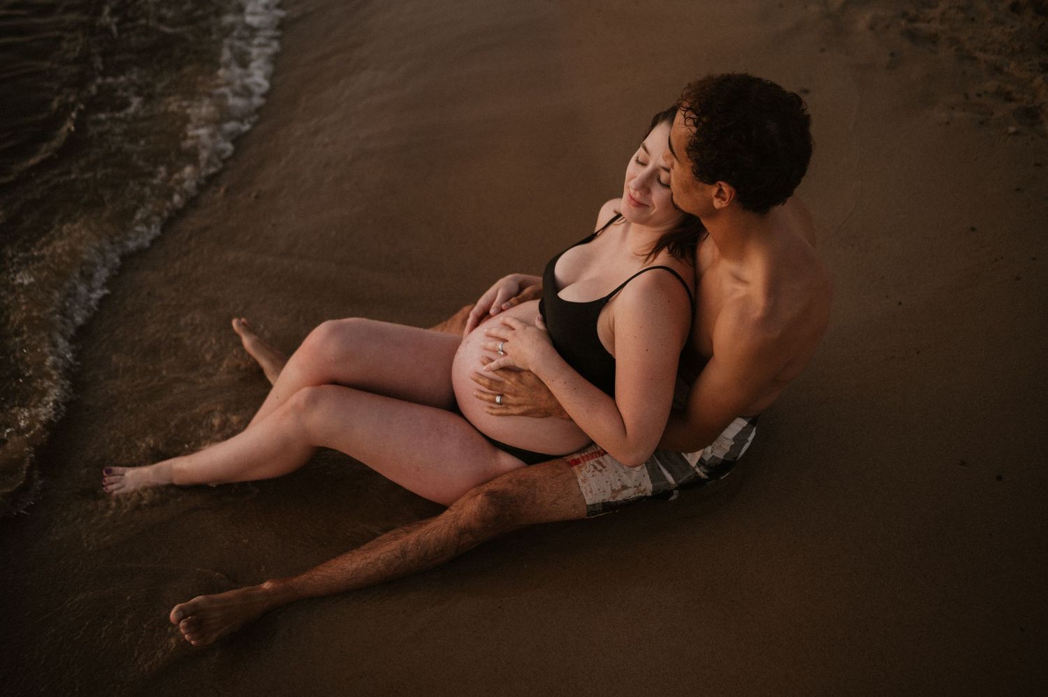 Romantic beach maternity photos with couple reclining on wet sand.