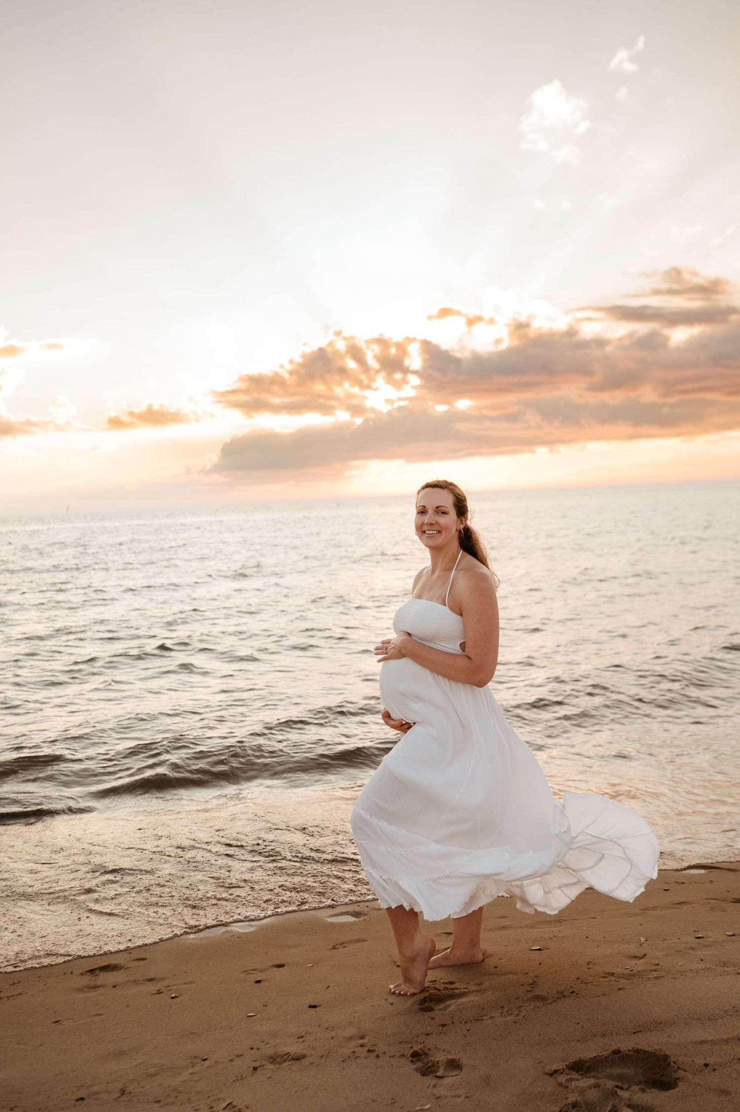 A white dress flows in the ocean breeze as someone poses on a beach at sunset with golden clouds in the sky.