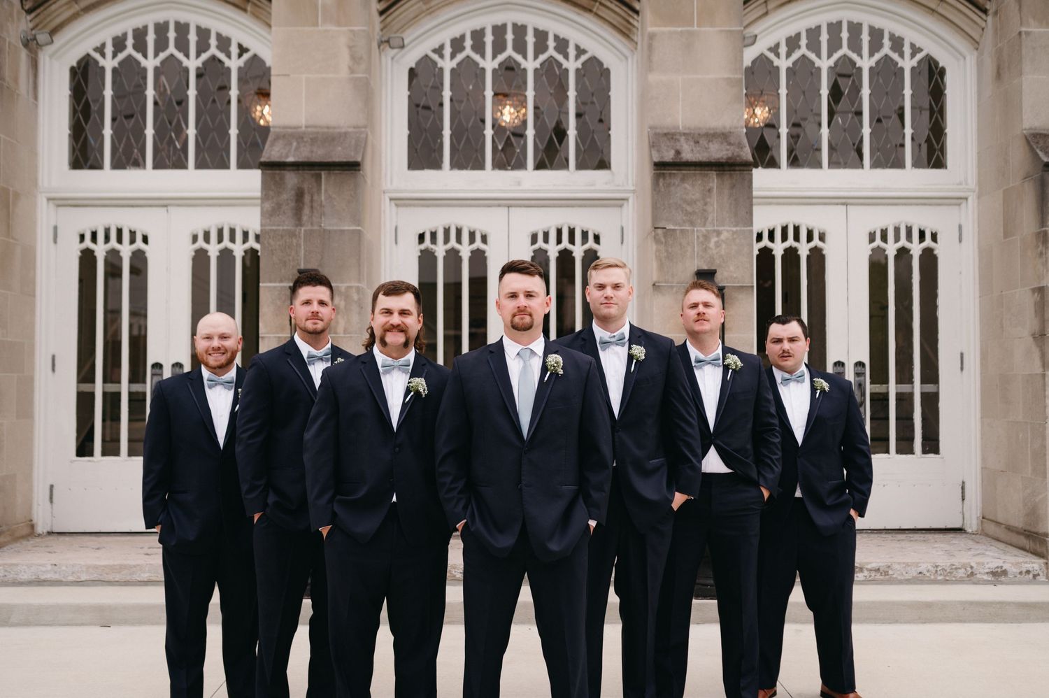Groomsmen in black tuxedos pose formally against gothic church windows.