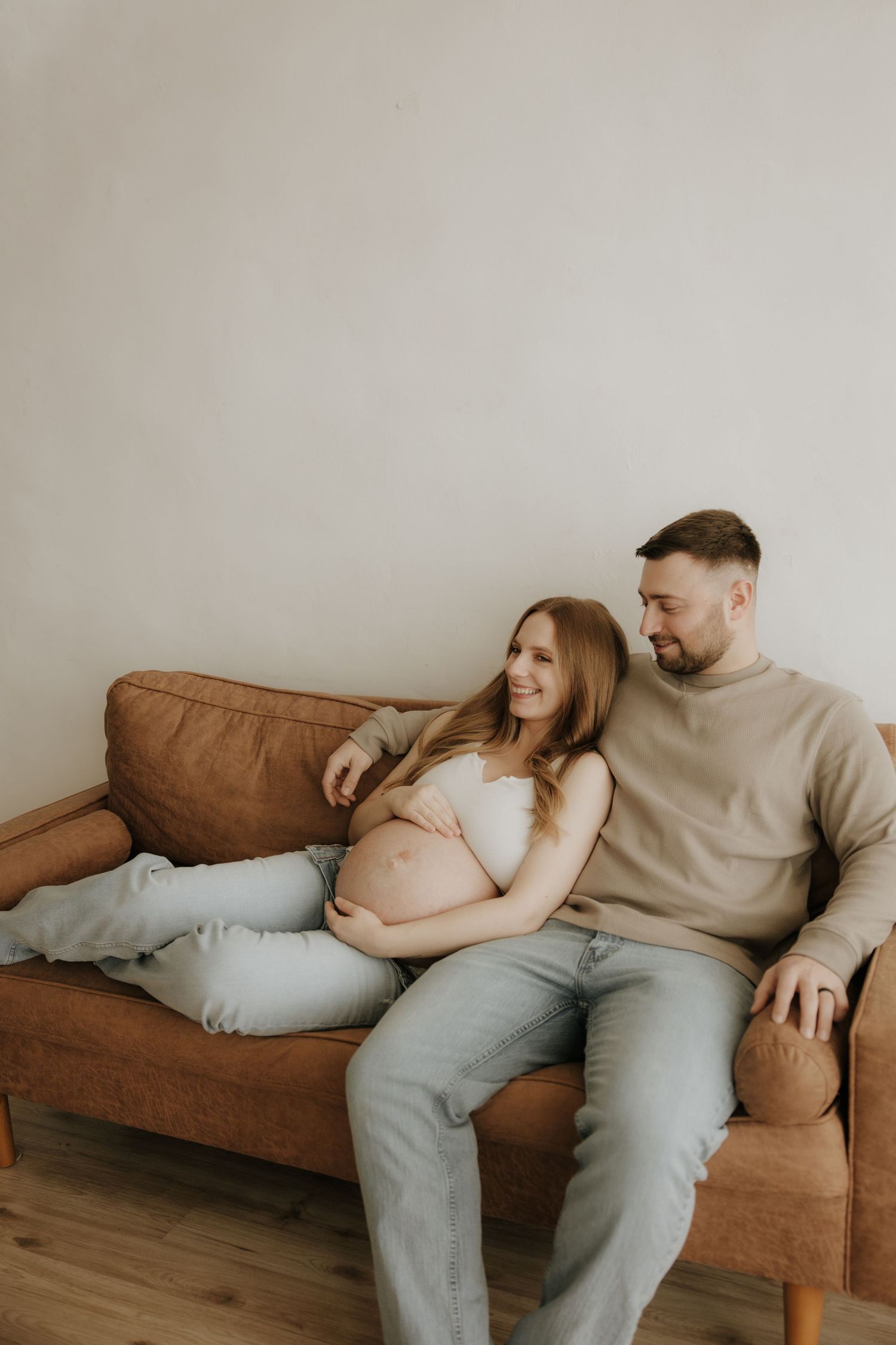 Candid maternity moment captured on brown leather sofa in natural window light.