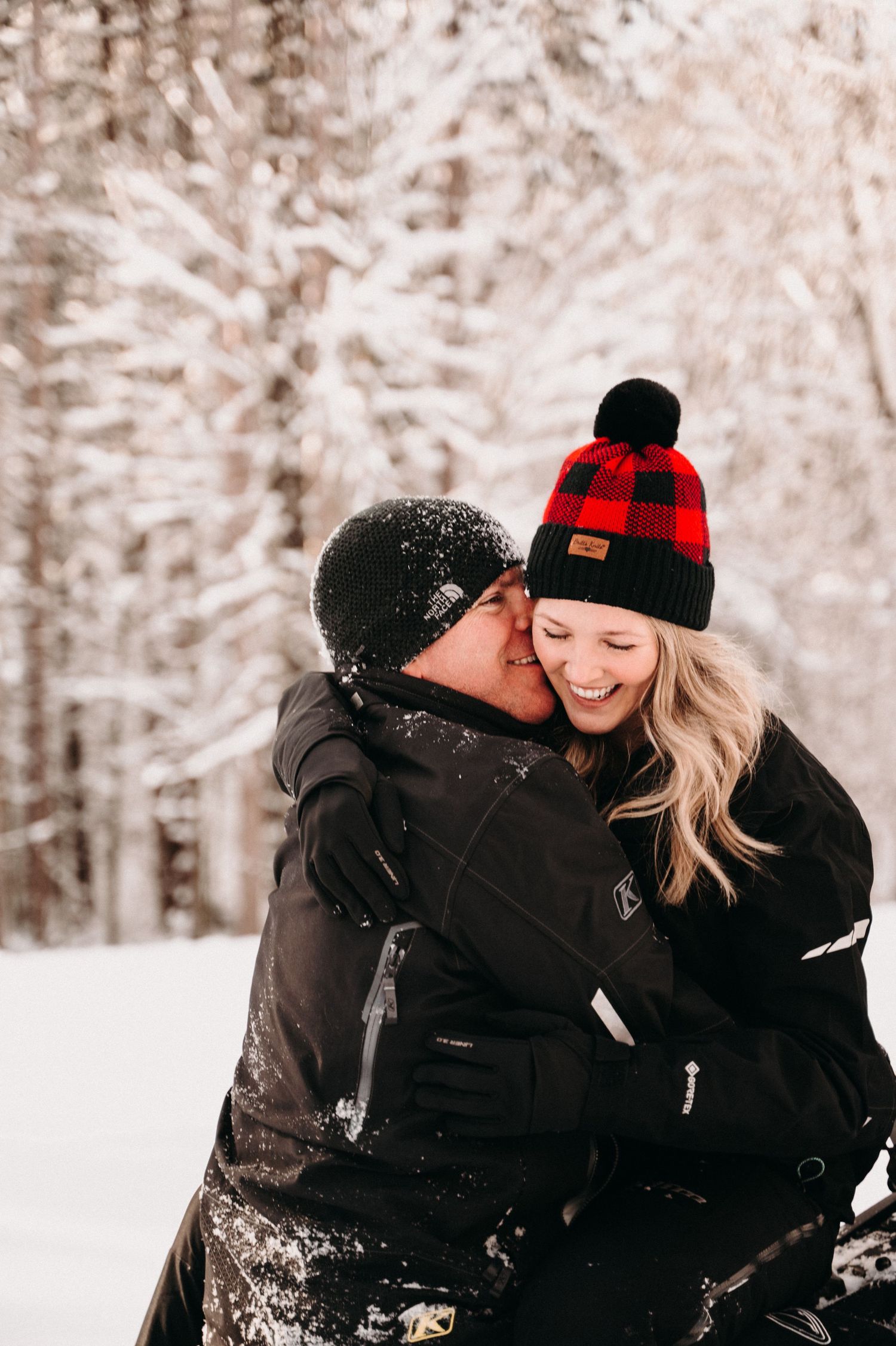 Couple in black winter coats sharing tender moment in snowy forest setting.