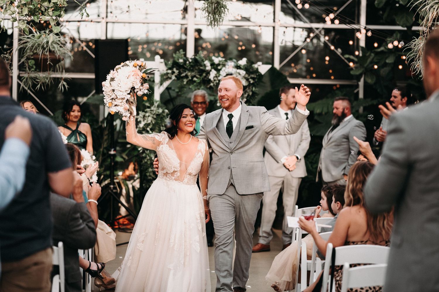 Joyful recessional walk down the aisle in greenhouse venue with string lights overhead.