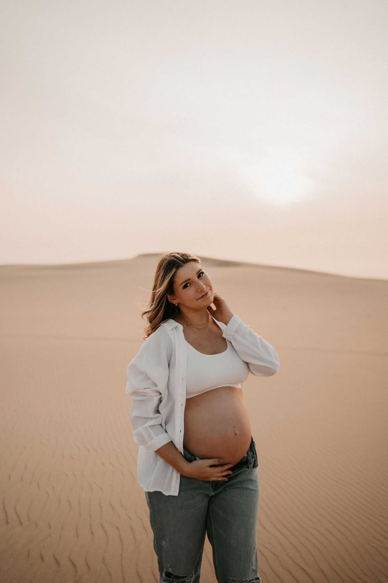 Pregnant woman in white top and jeans poses thoughtfully in desert landscape.
