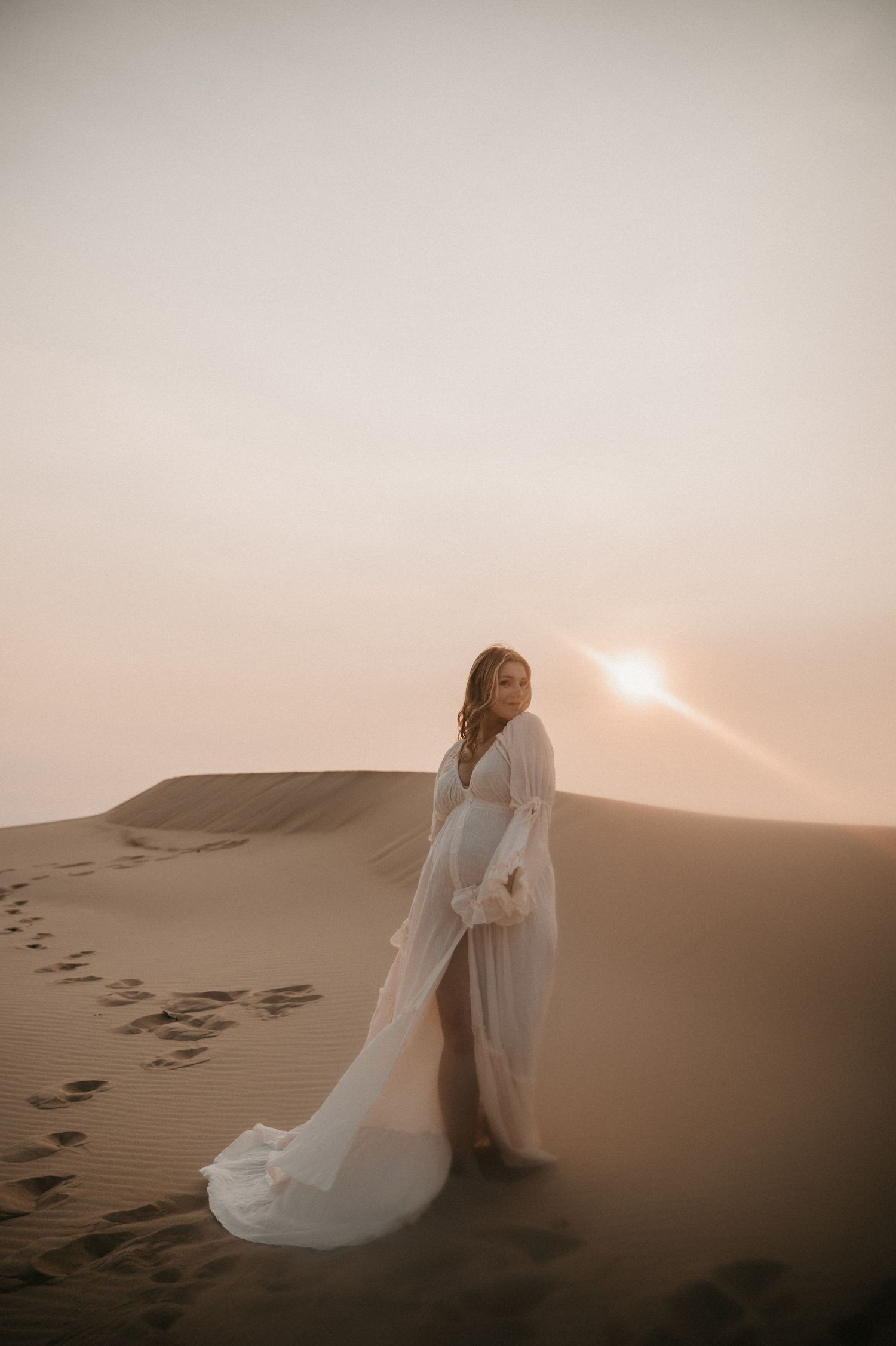 A long white dress trails behind a silhouette standing on desert dunes during golden hour.