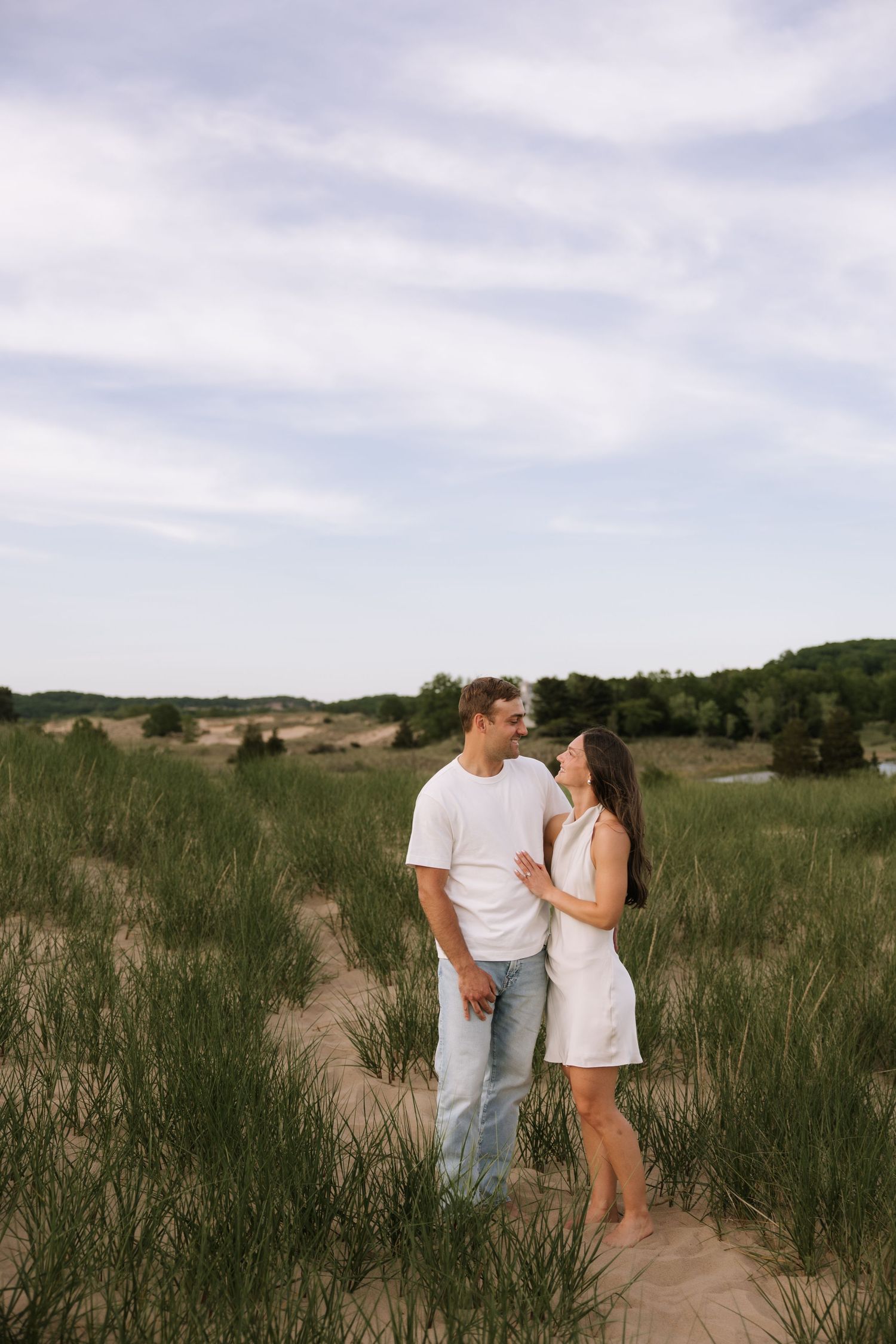 Couple in white outfits standing together in sandy dunes with green grass and trees in background.