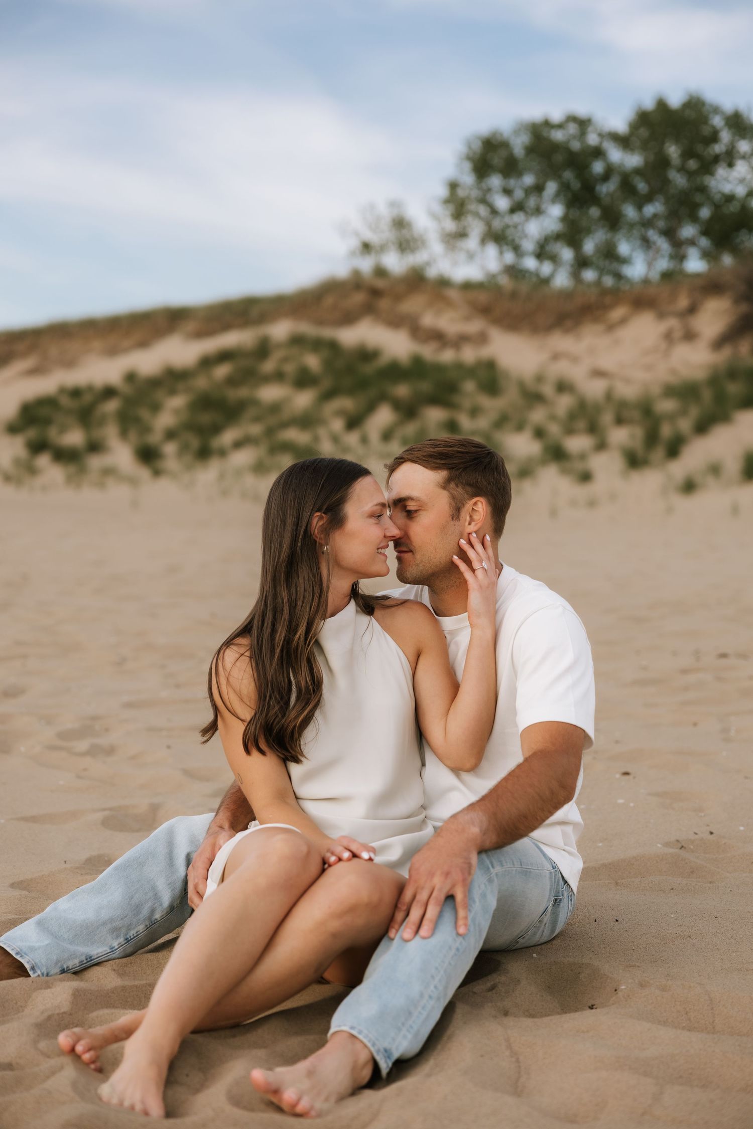 Couple sitting close together on sandy beach dunes, sharing a romantic moment in casual white tops and jeans.