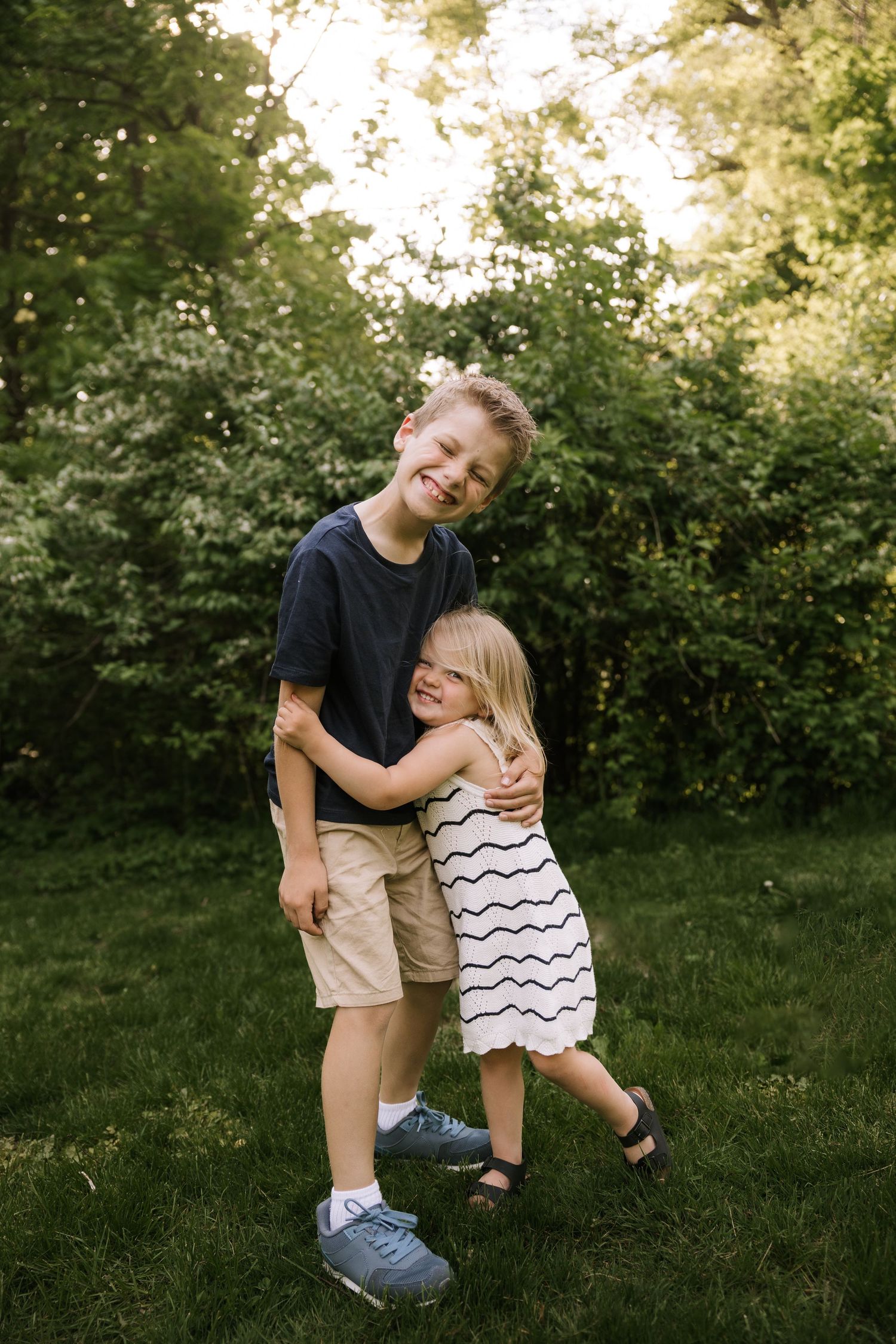 Two young siblings hugging and smiling together on green grass with trees in background.