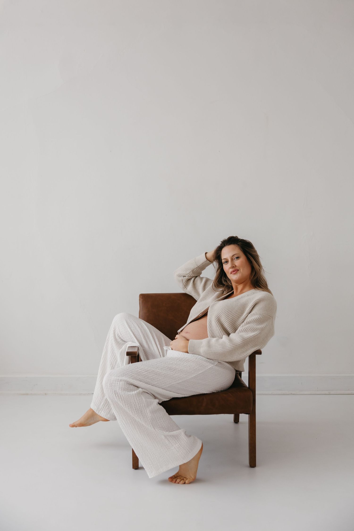 Pregnant woman in beige cardigan sitting relaxed in brown leather chair on white background.