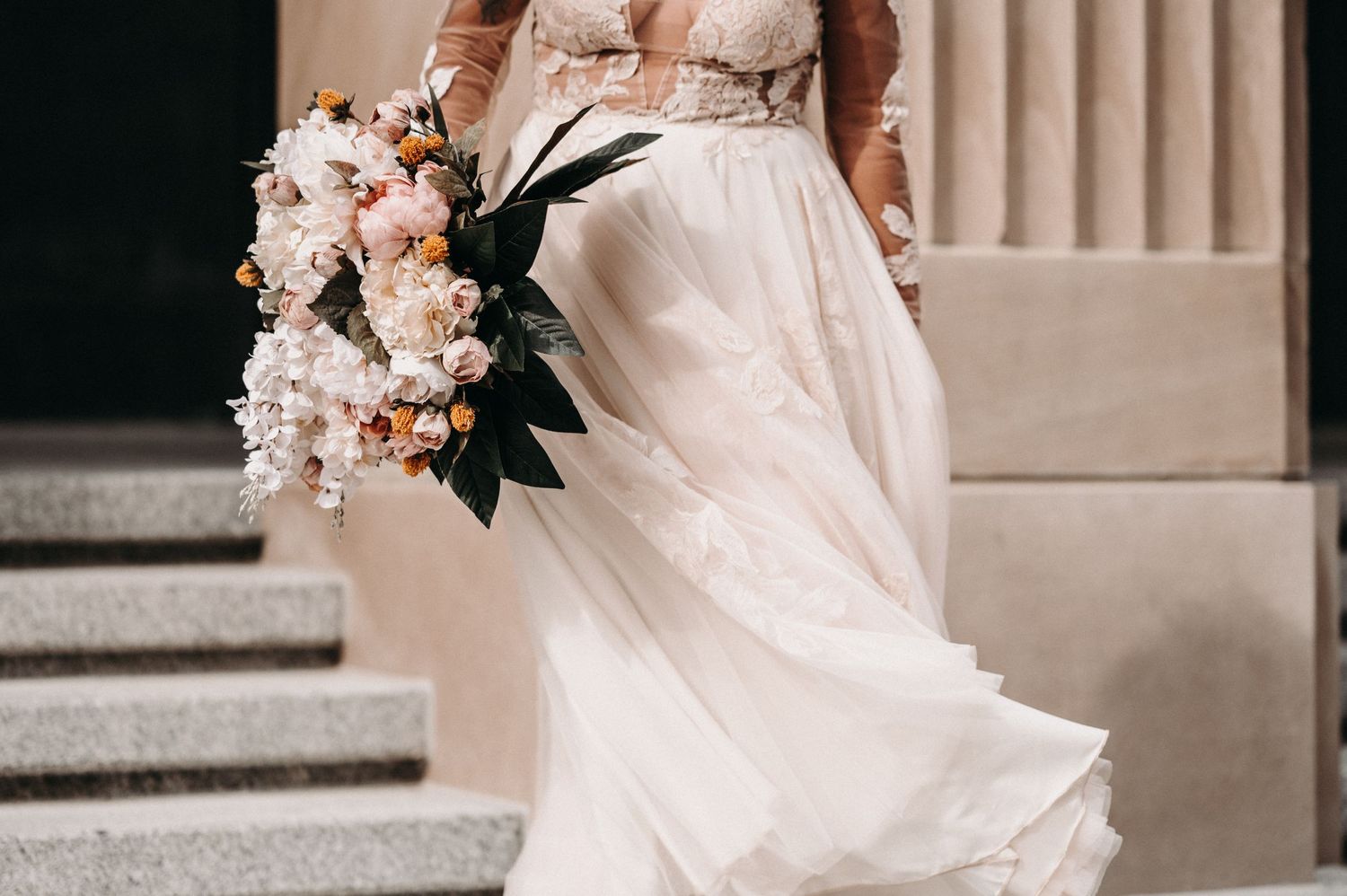 A close-up view of a flowing white wedding dress and pink floral bouquet against stone columns.
