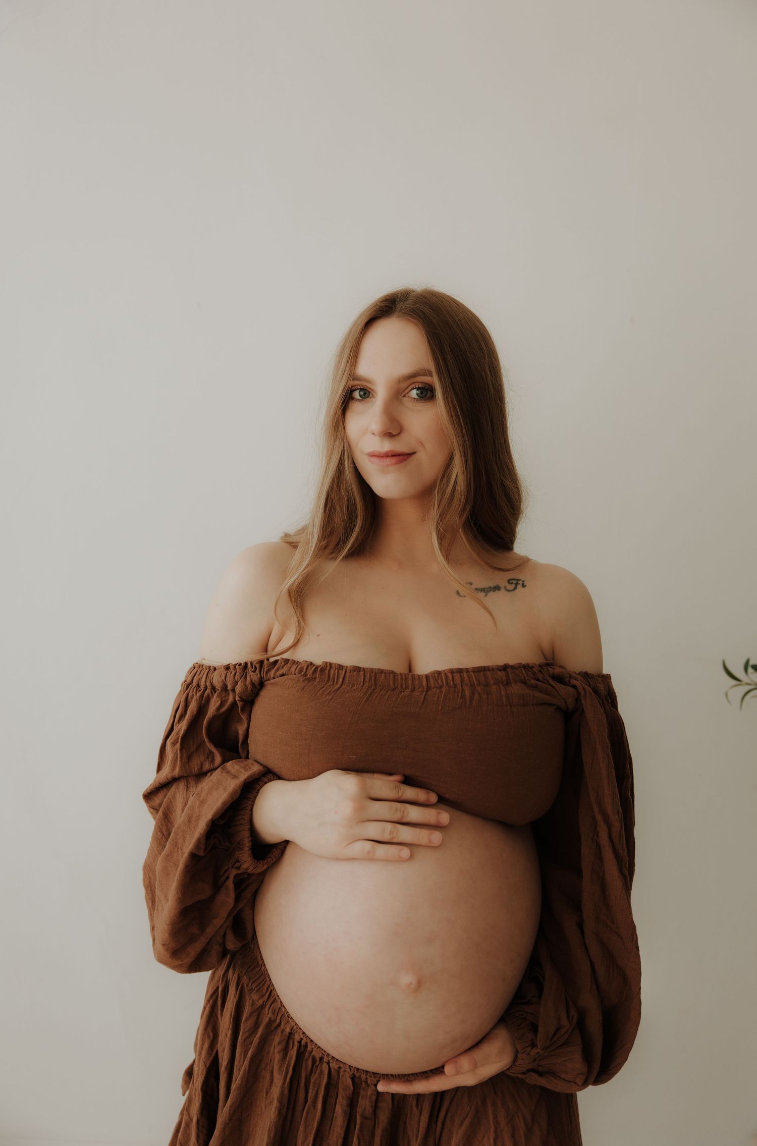 Maternity photos in brown off-shoulder dress highlighting pregnancy against white wall.