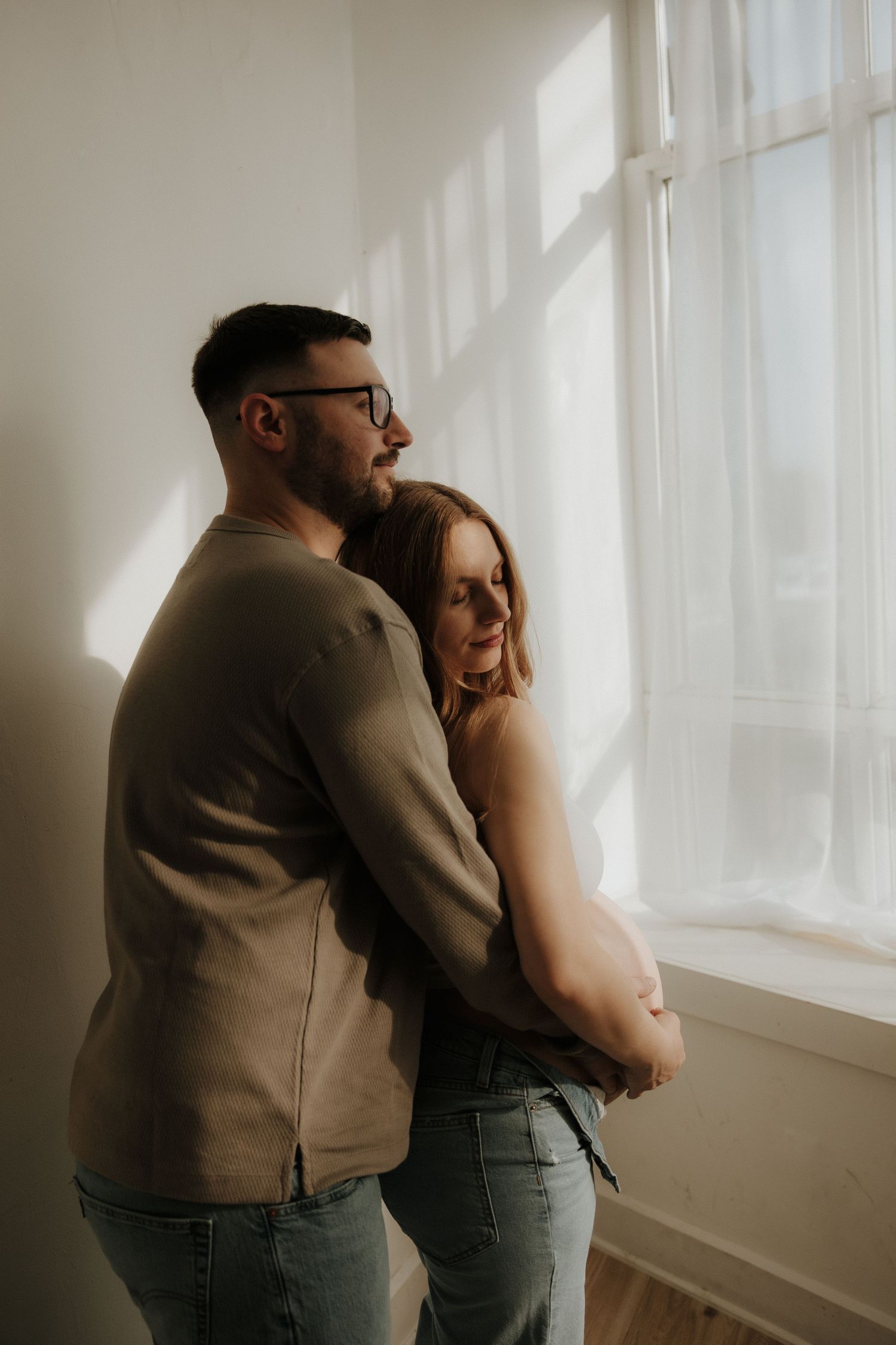 Minimalist maternity photos showing tender moments between expectant parents near a bright window.
