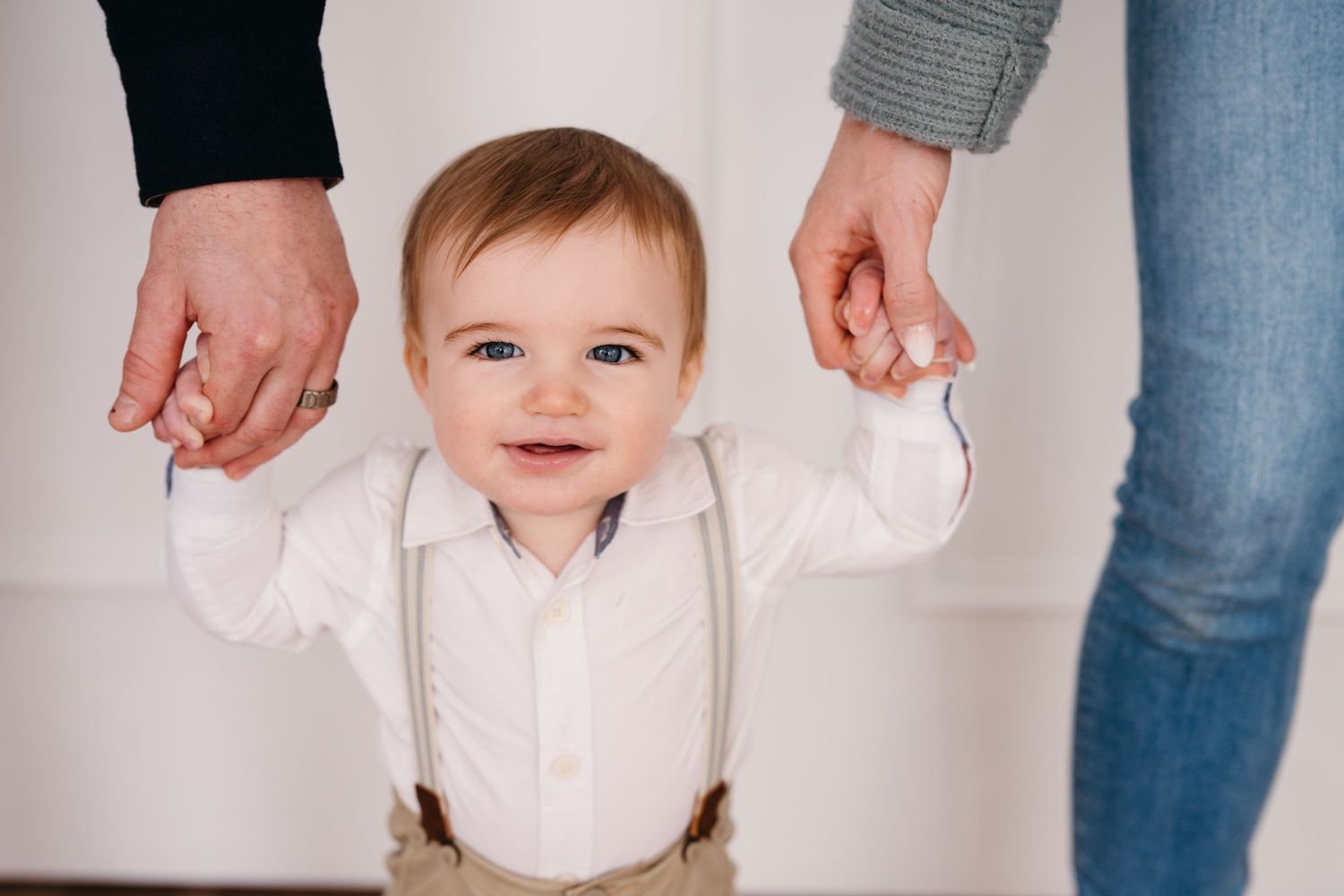 Baby in white shirt and suspenders holds hands with adults while learning to walk against white wall.