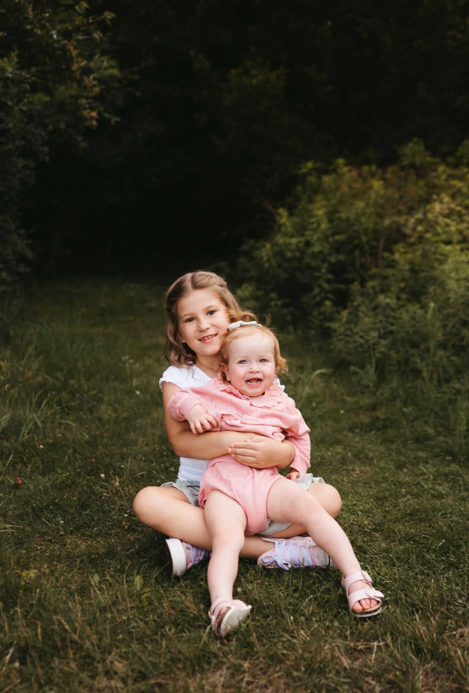 Two children in casual summer outfits sit together on grassy ground in a natural outdoor setting.